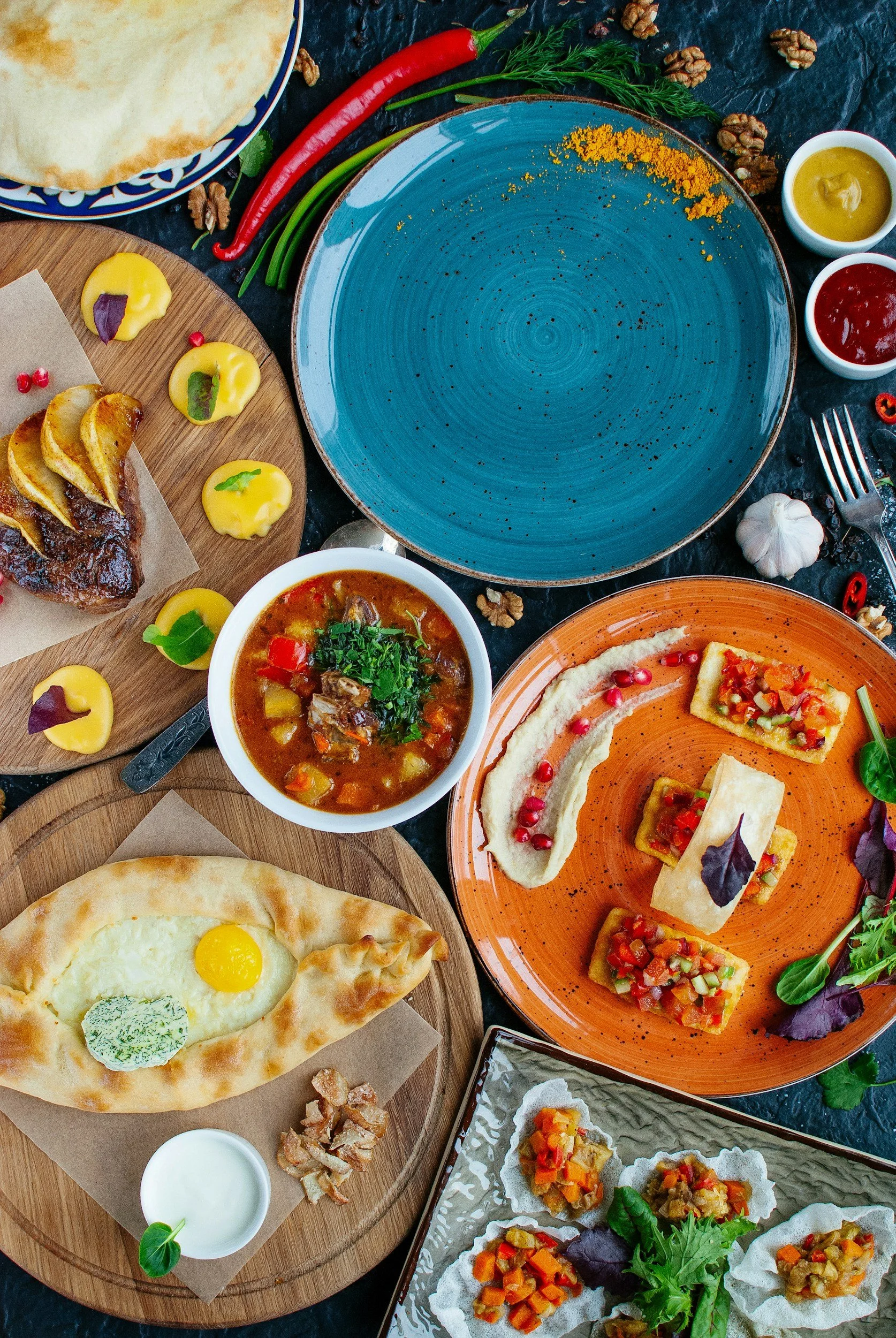 A colorful spread of Middle Eastern dishes including hummus, pita bread with a fried egg and herb butter, a meat and vegetable stew, baked flatbread with toppings, stuffed pastries, and various dipping sauces, garnished with herbs and vegetables.