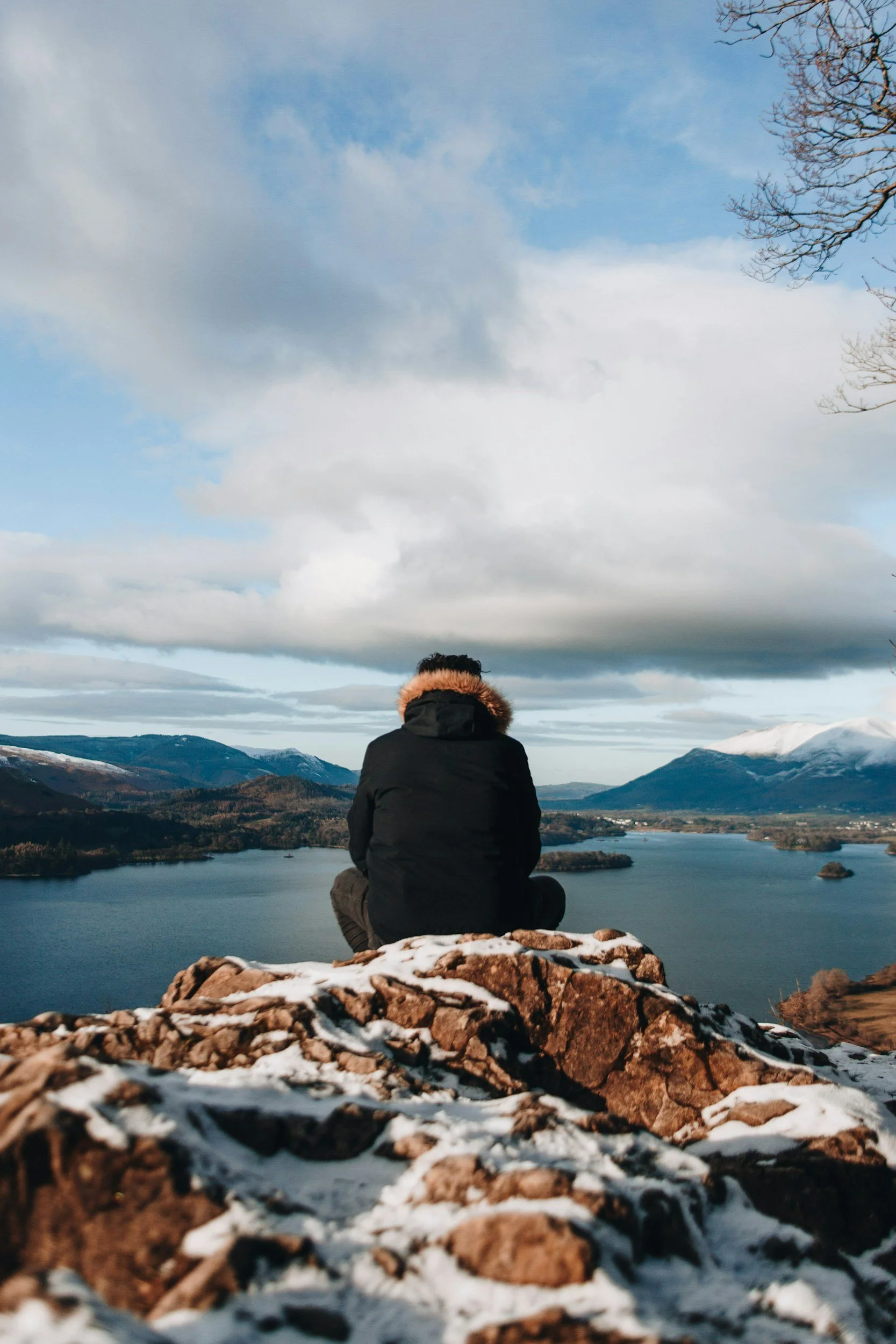Person sitting on a snow-dusted rock ledge overlooking a lake with mountains in the distance under a cloudy sky