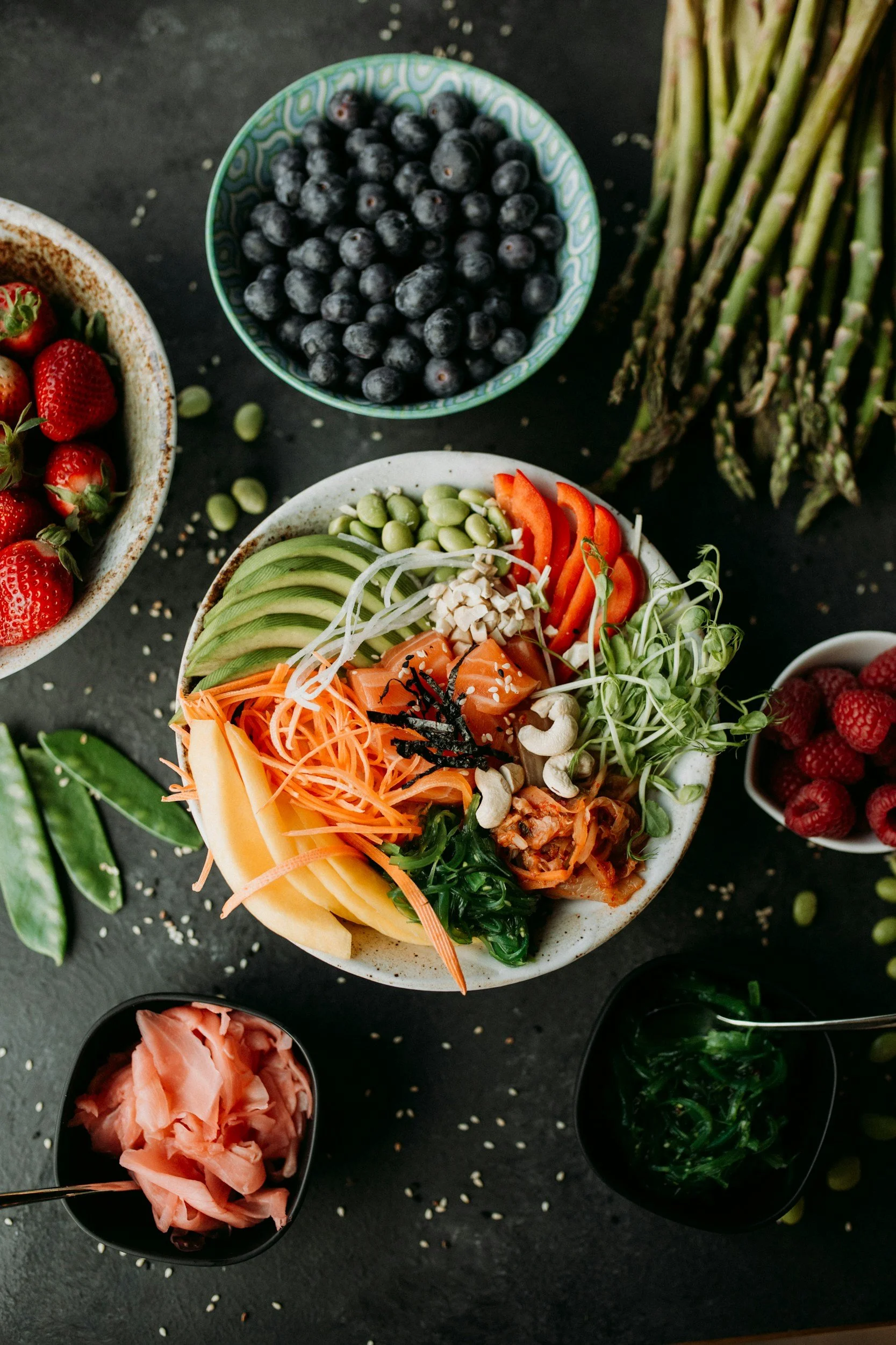 A bowl of assorted fresh vegetables and fruits, including carrots, avocado, cucumber, and radish, surrounded by bowls of blueberries, strawberries, raspberries, and snow peas on a dark surface.