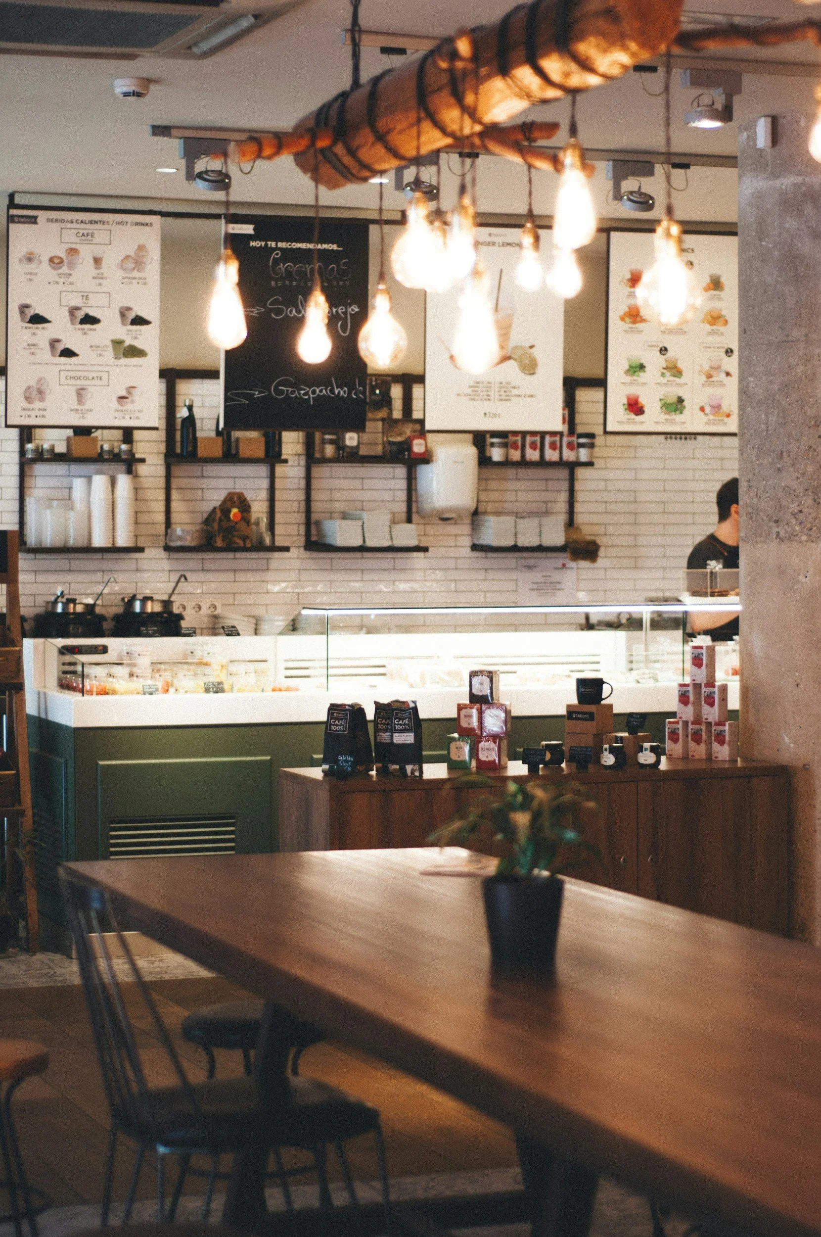Inside a cozy coffee shop with a wooden table and a potted plant. The background shows a counter with a glass display case, menu boards on the wall, and hanging lights above.