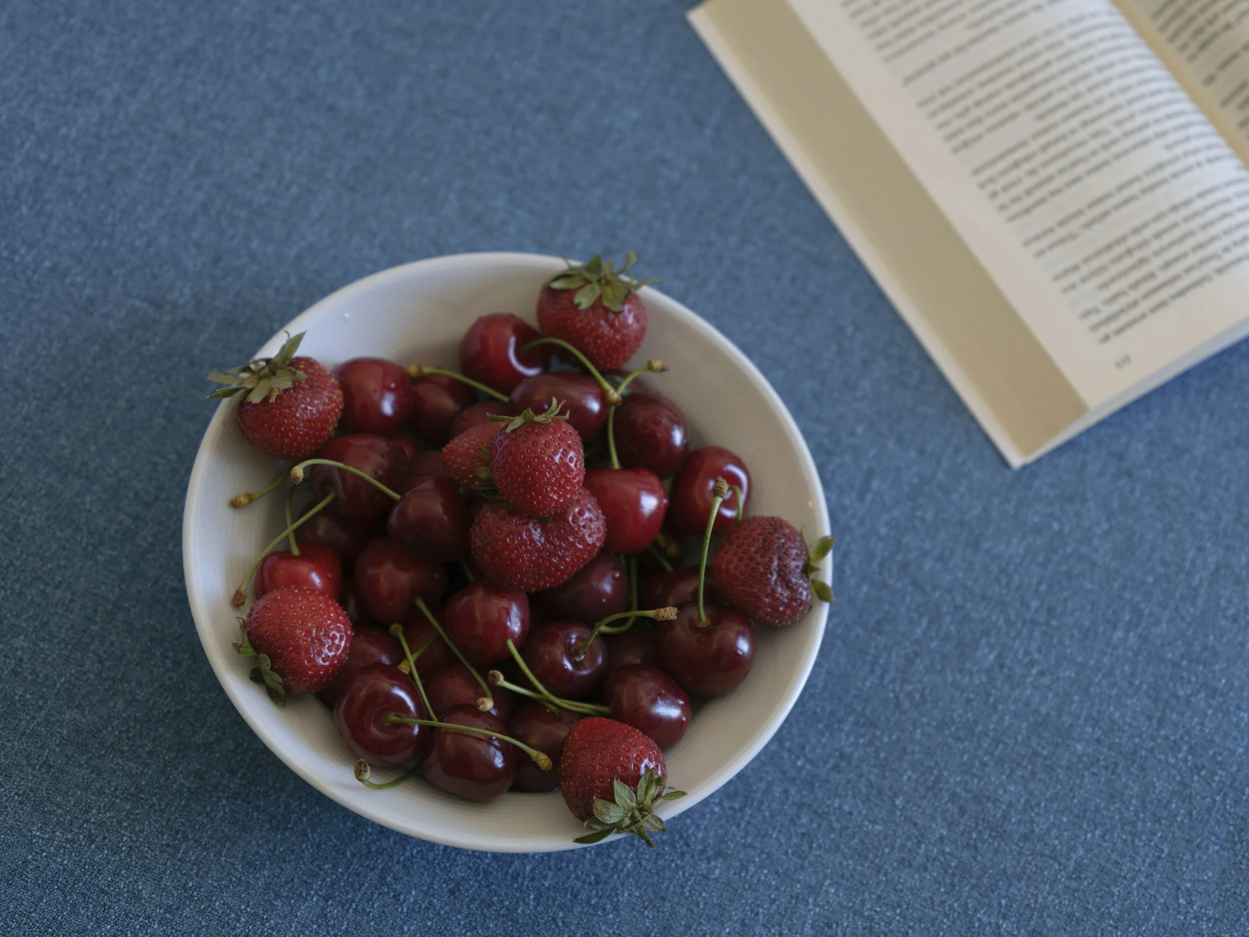 A white bowl filled with fresh red cherries and strawberries placed on a blue fabric surface. An open book lies nearby.