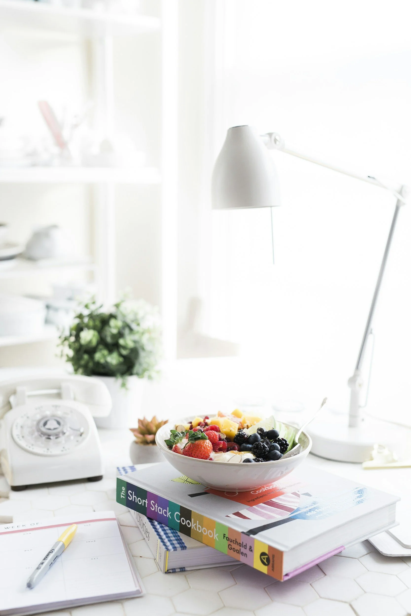 A white bowl filled with assorted fresh fruits, including strawberries, blueberries, raspberries, and chopped pineapple, placed on top of colorful cookbooks on a white kitchen counter. In the background, there is a vintage white rotary telephone, a pen on an open planner, a small potted plant, and a modern white desk lamp near a window with bright natural light.