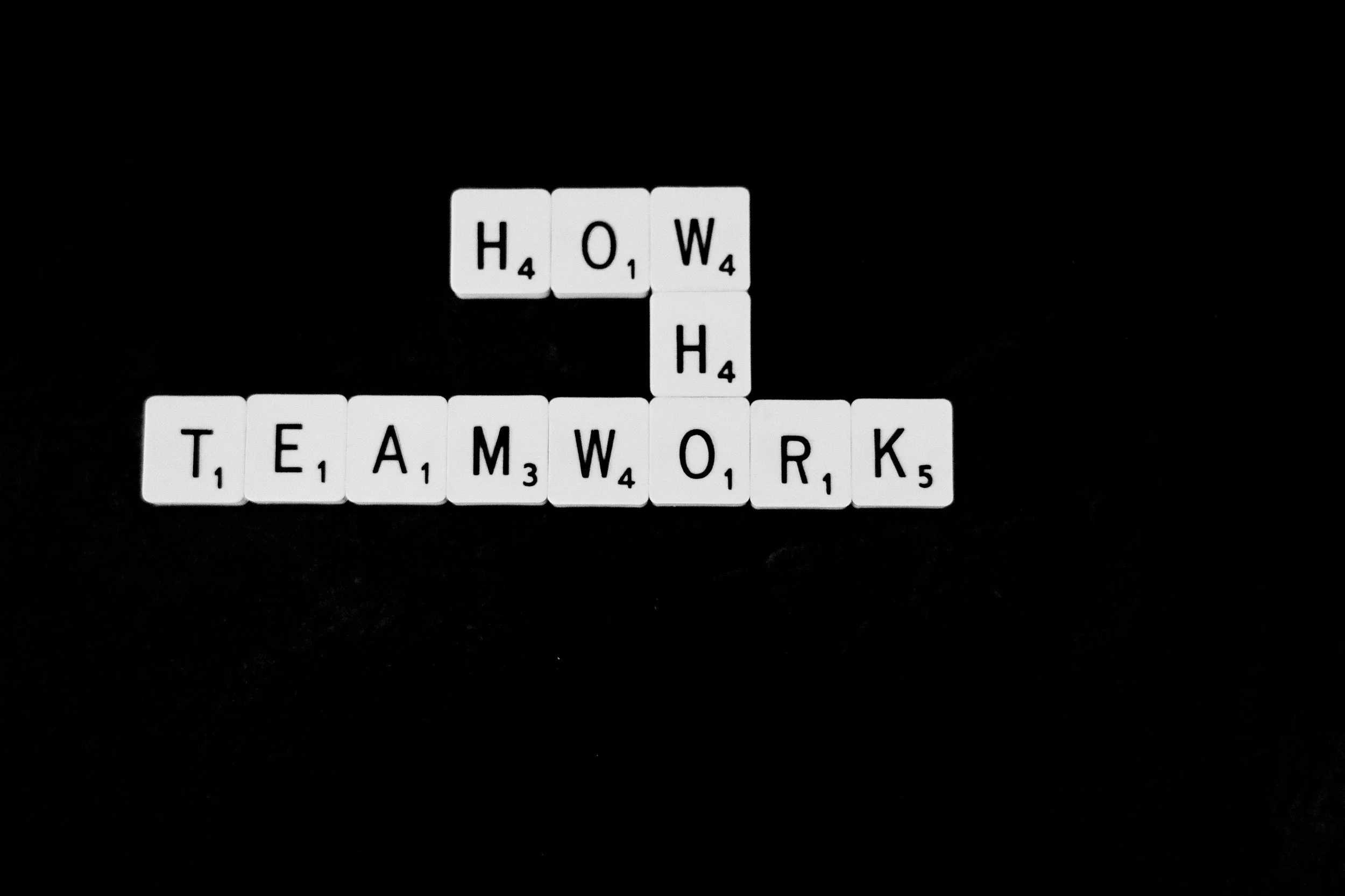 Scrabble tiles spelling 'HOW' above 'TEAMWORK' on a black background.