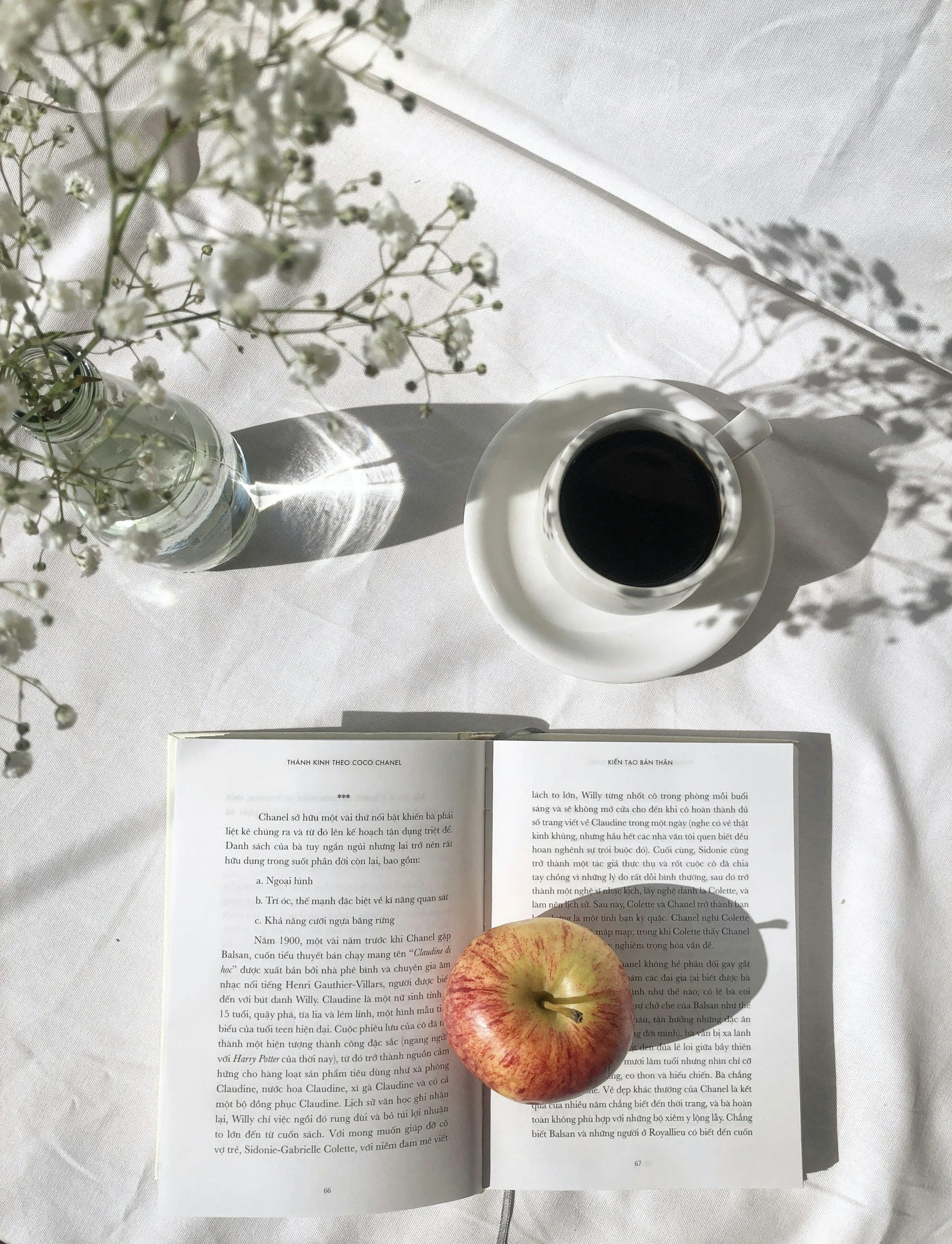 A top view of a table with a vase of white flowers, a cup of black coffee on a saucer, an open book, and a red and yellow apple placed on the open book. The table is covered with a white cloth, and sunlight creates shadows of the flowers on the table.
