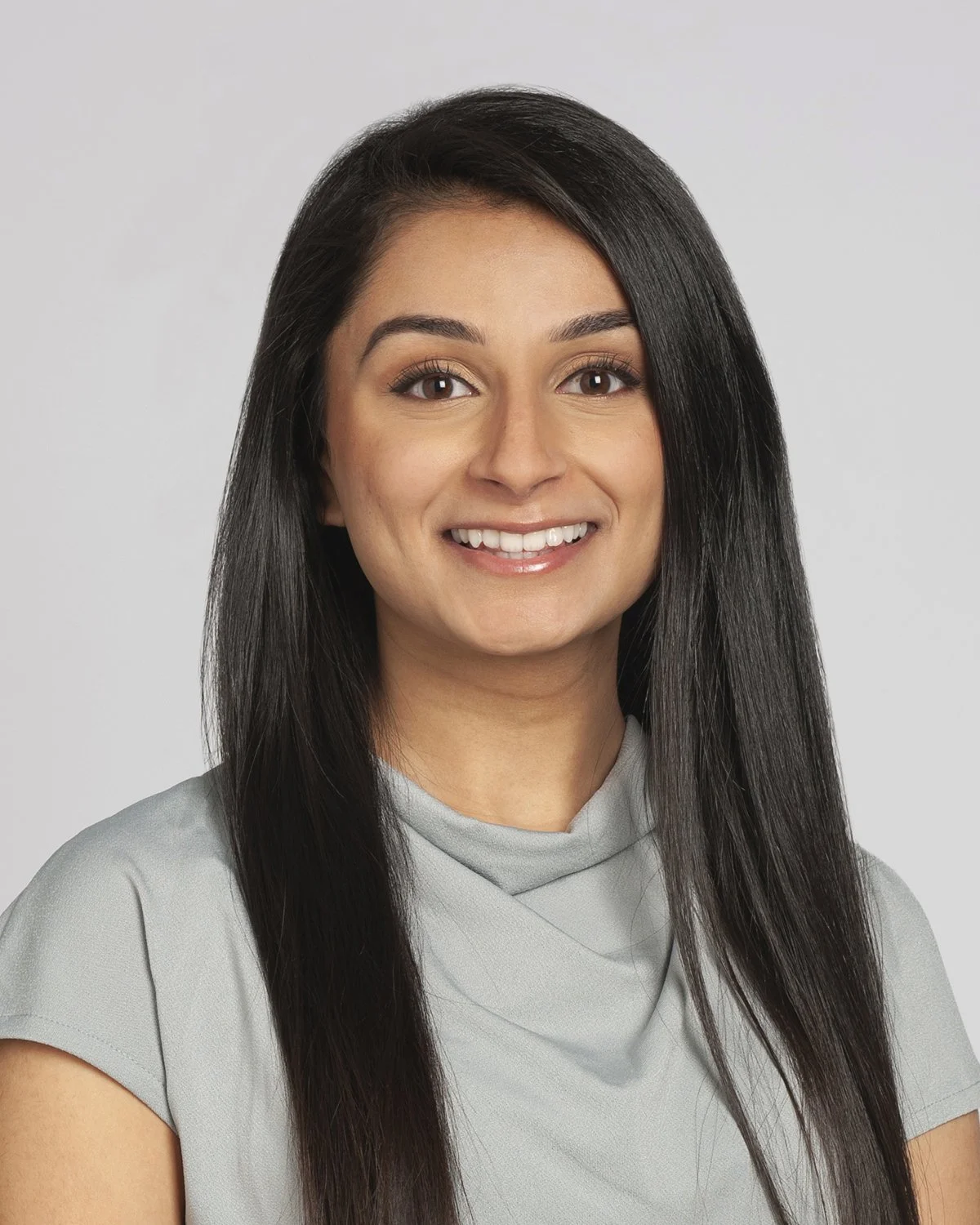 Headshot of a woman with long black hair, smiling, wearing a light gray top, against a plain gray background.