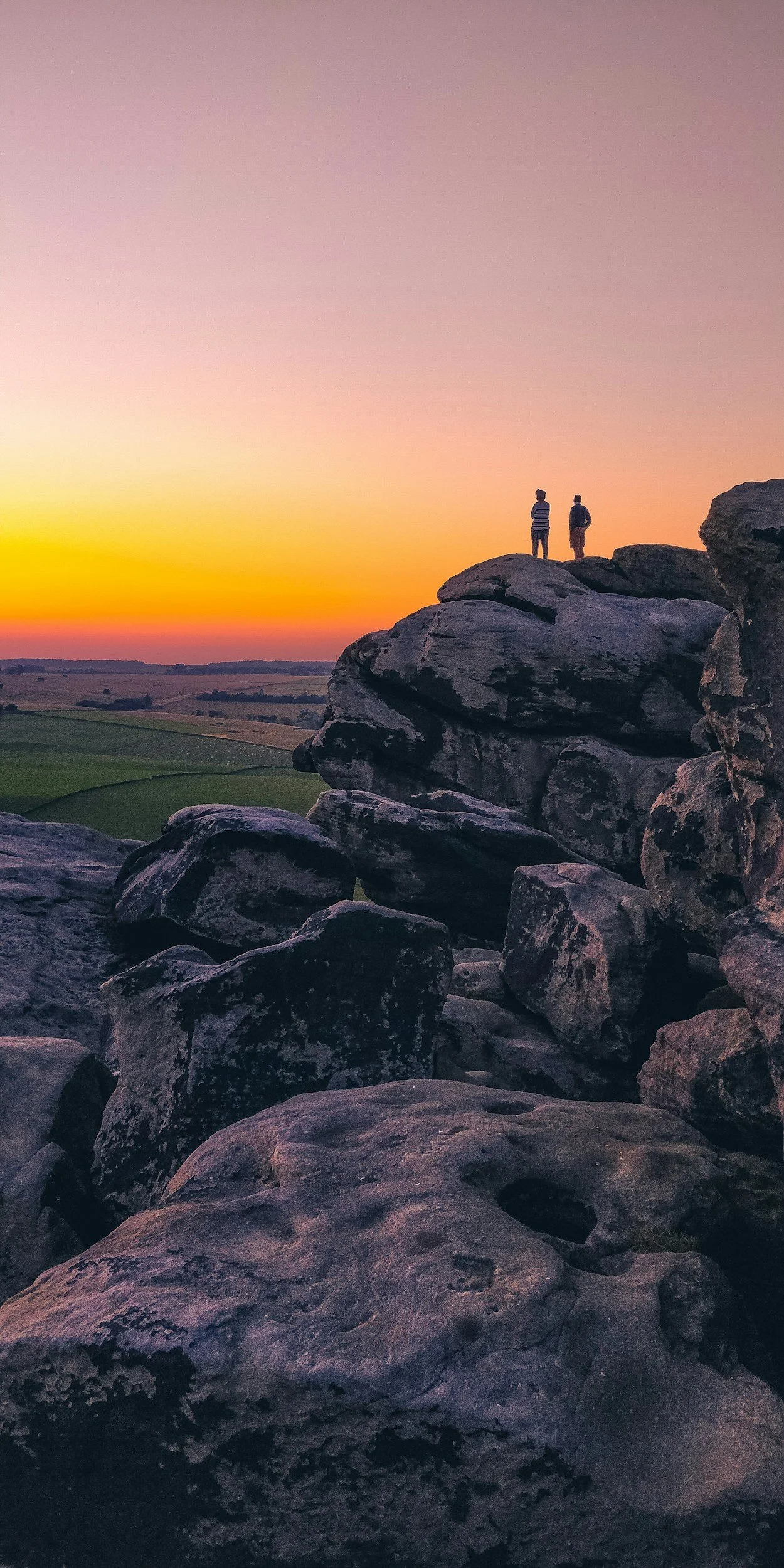 Two people standing on large rock formations at sunset with a colorful sky and open landscape in the background.