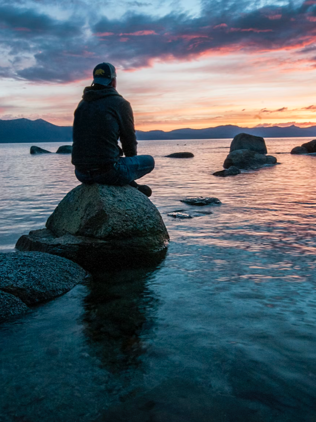 A person sitting on a large rock at the edge of a body of water during sunset, with colorful sky and distant mountains in the background.
