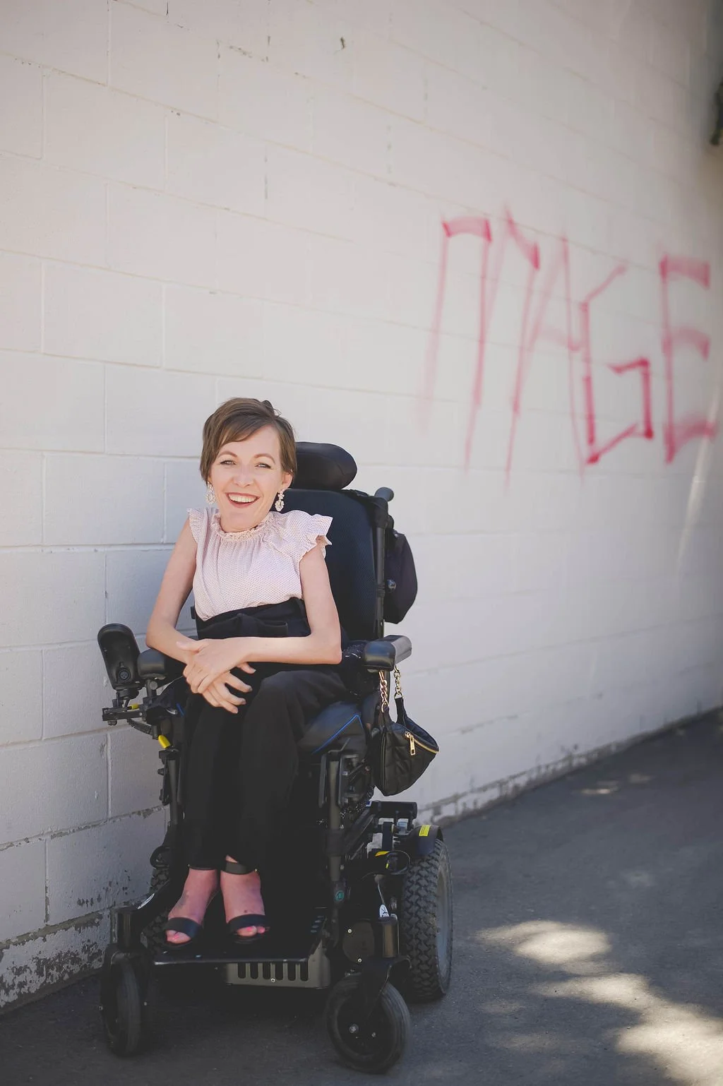 A woman with short hair smiling while sitting in a motorized wheelchair, standing against a white brick wall with red graffiti.
