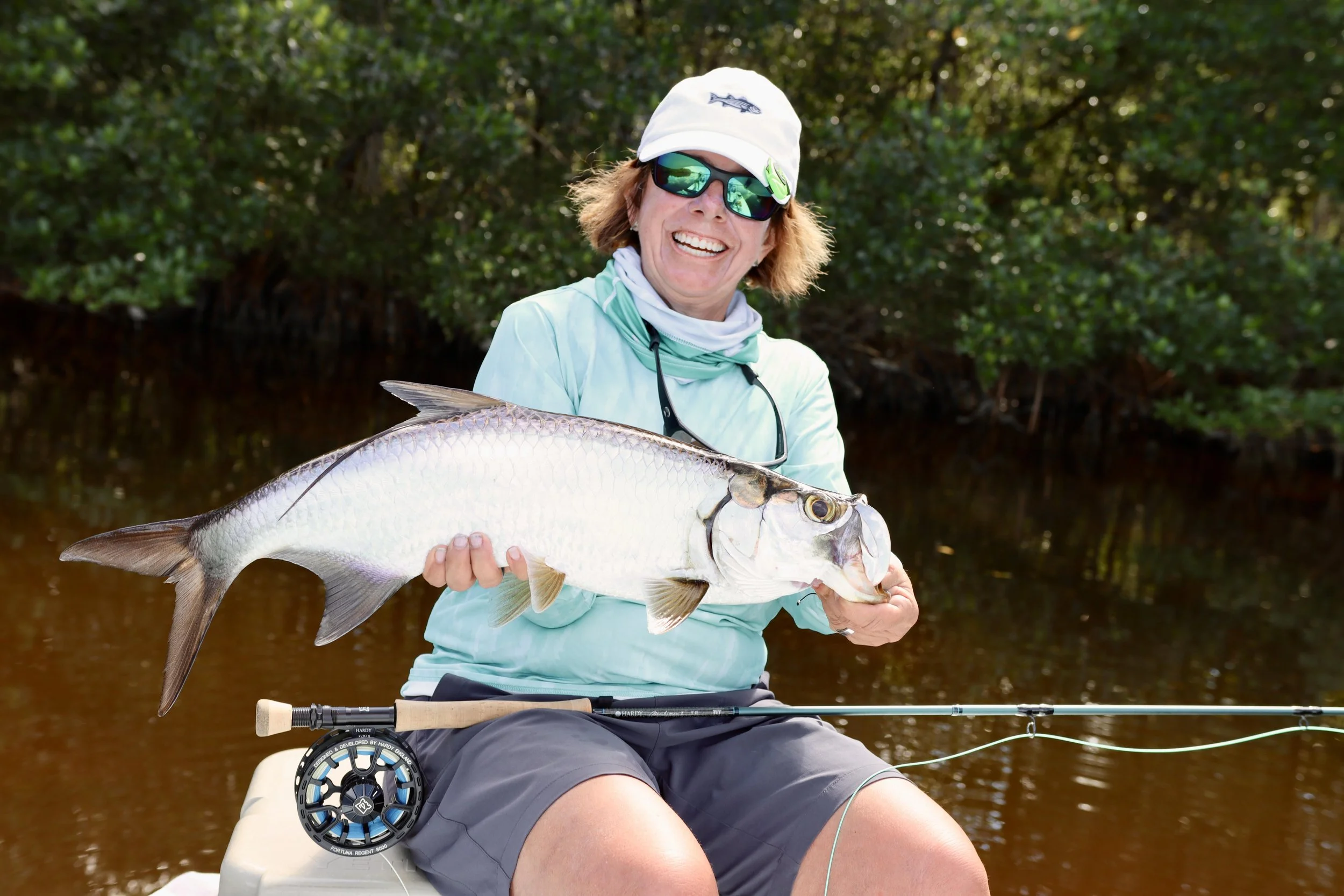 Woman in sunglasses, a white hat, and outdoor fishing attire sitting in a boat while holding a large fish. The background features water and dense green foliage.
