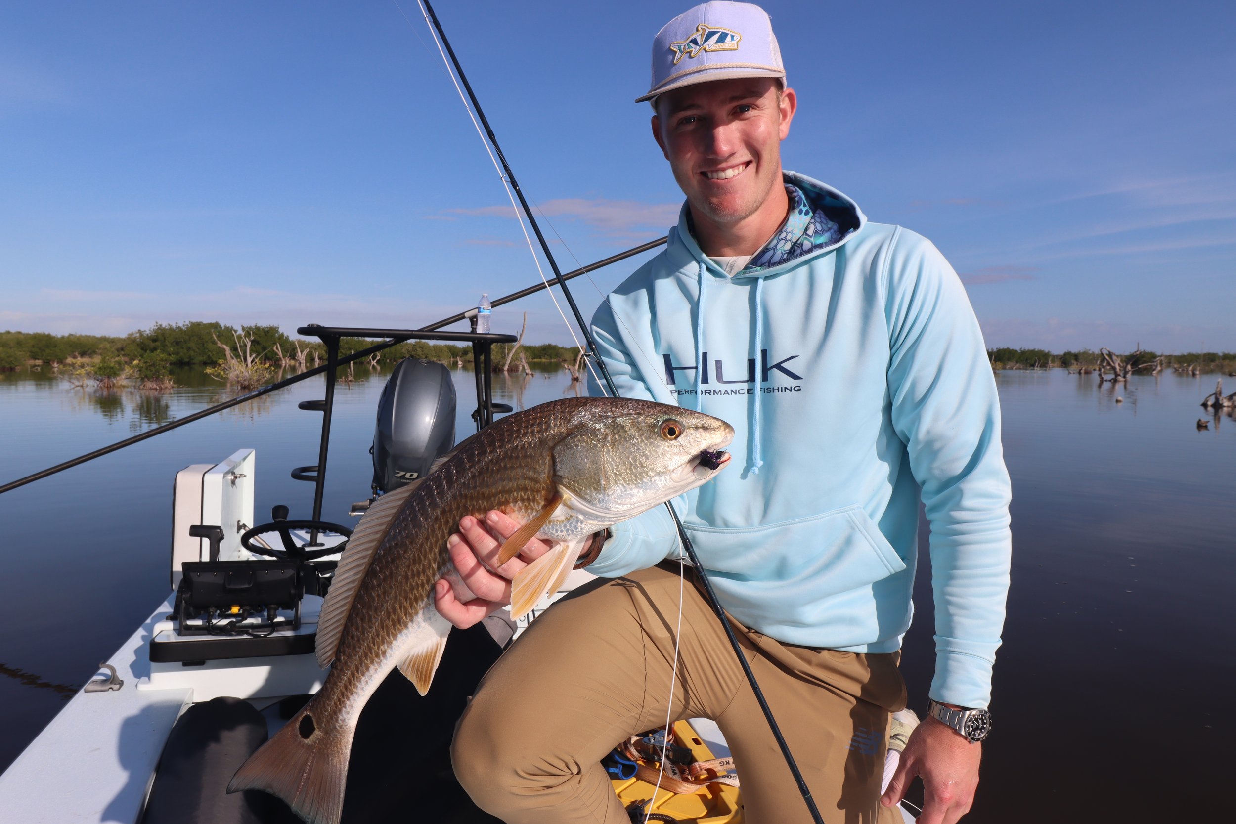 A young man kneeling on a boat holding a large fish he caught, with water and trees in the background. He's smiling and wearing a light blue hoodie and a gray cap.