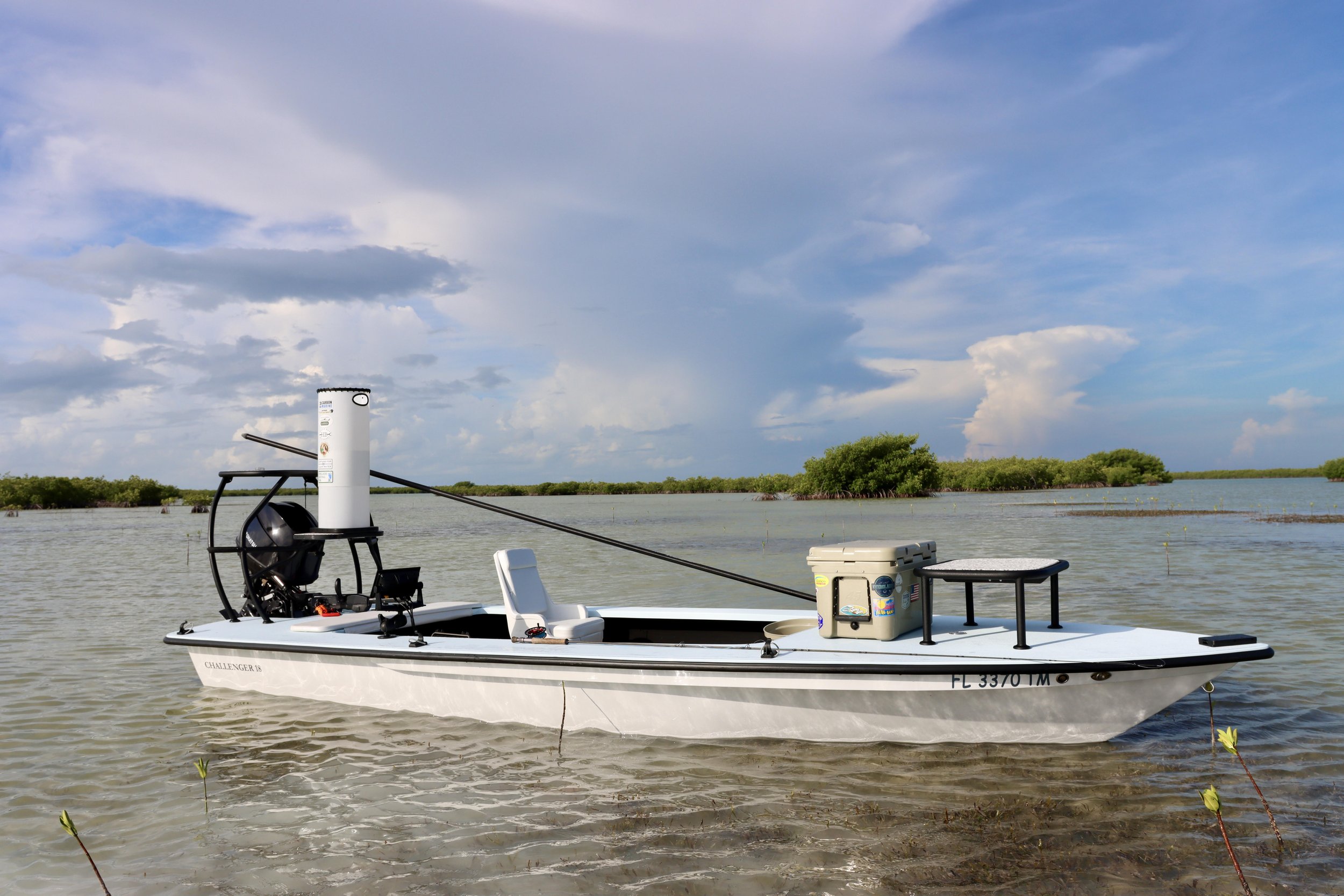Small boat in shallow water with a seat, cooler, and motor, with small islands of trees in the background under a partly cloudy sky.