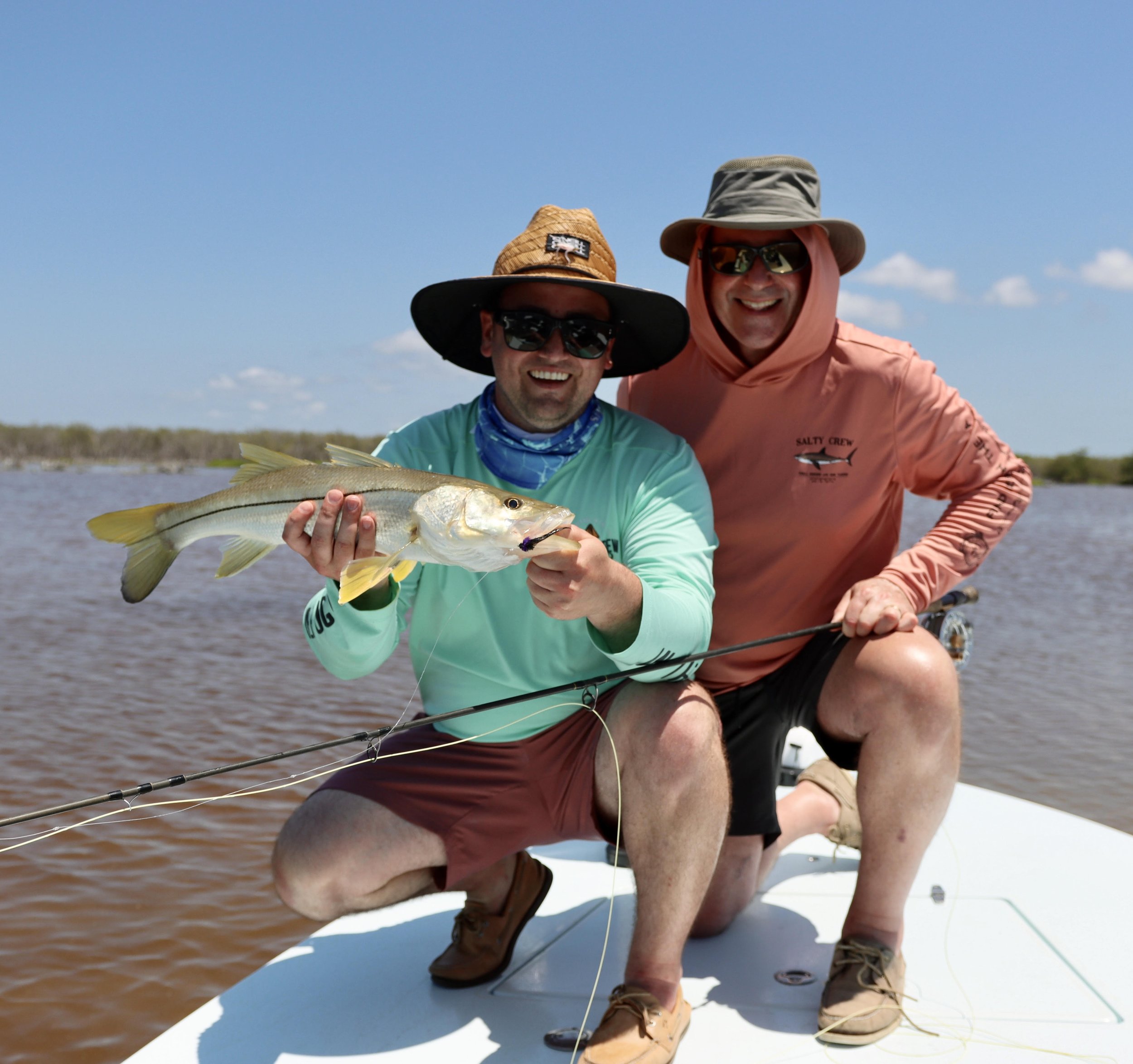 Two men on a boat in a waterway during daytime, one holding a fish they caught, smiling at the camera, both wearing hats and sunglasses, with a background of water and distant shoreline.