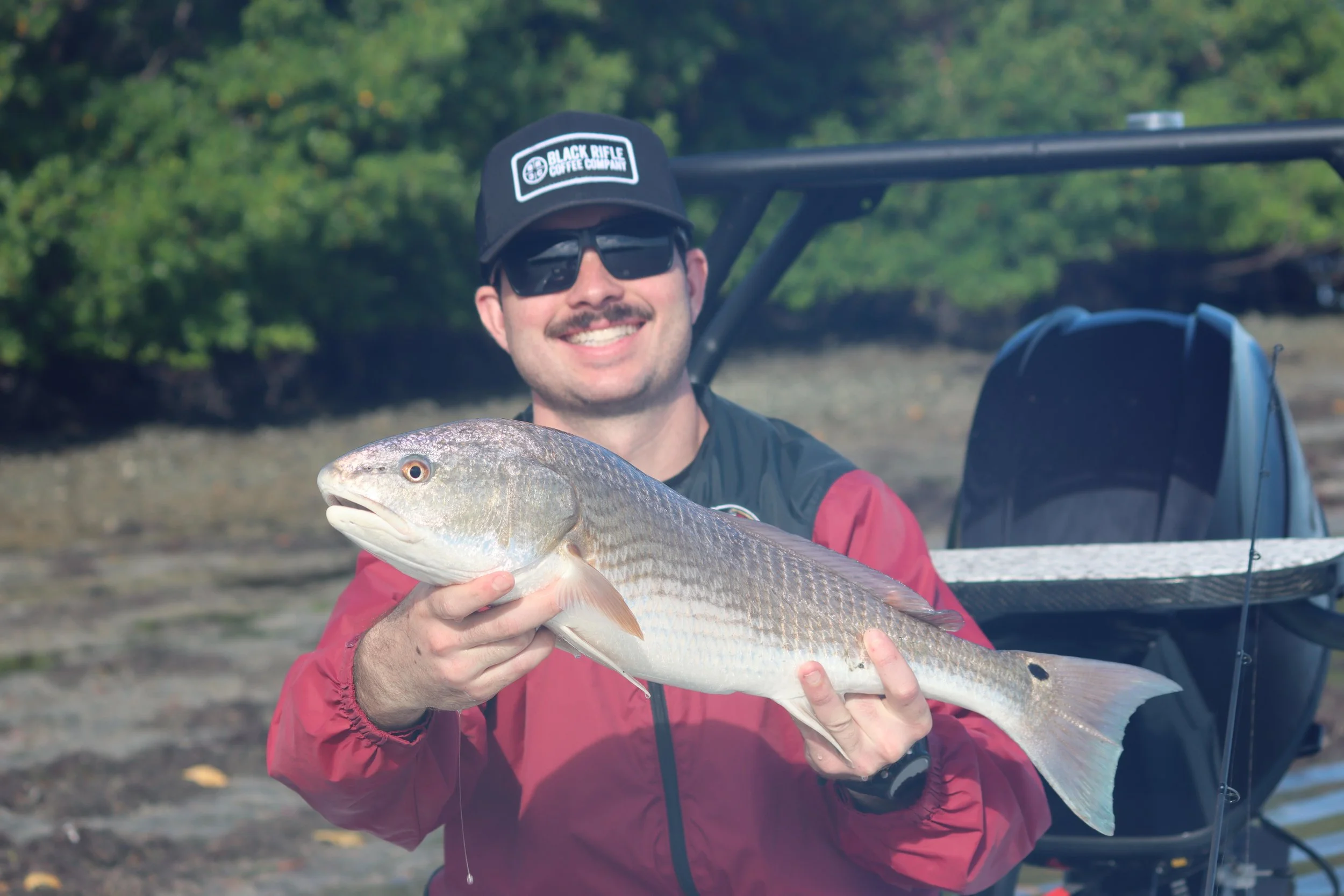 A man smiling and wearing sunglasses and a cap with a logo, holding a fish he caught during fishing outdoors near a wooded area.
