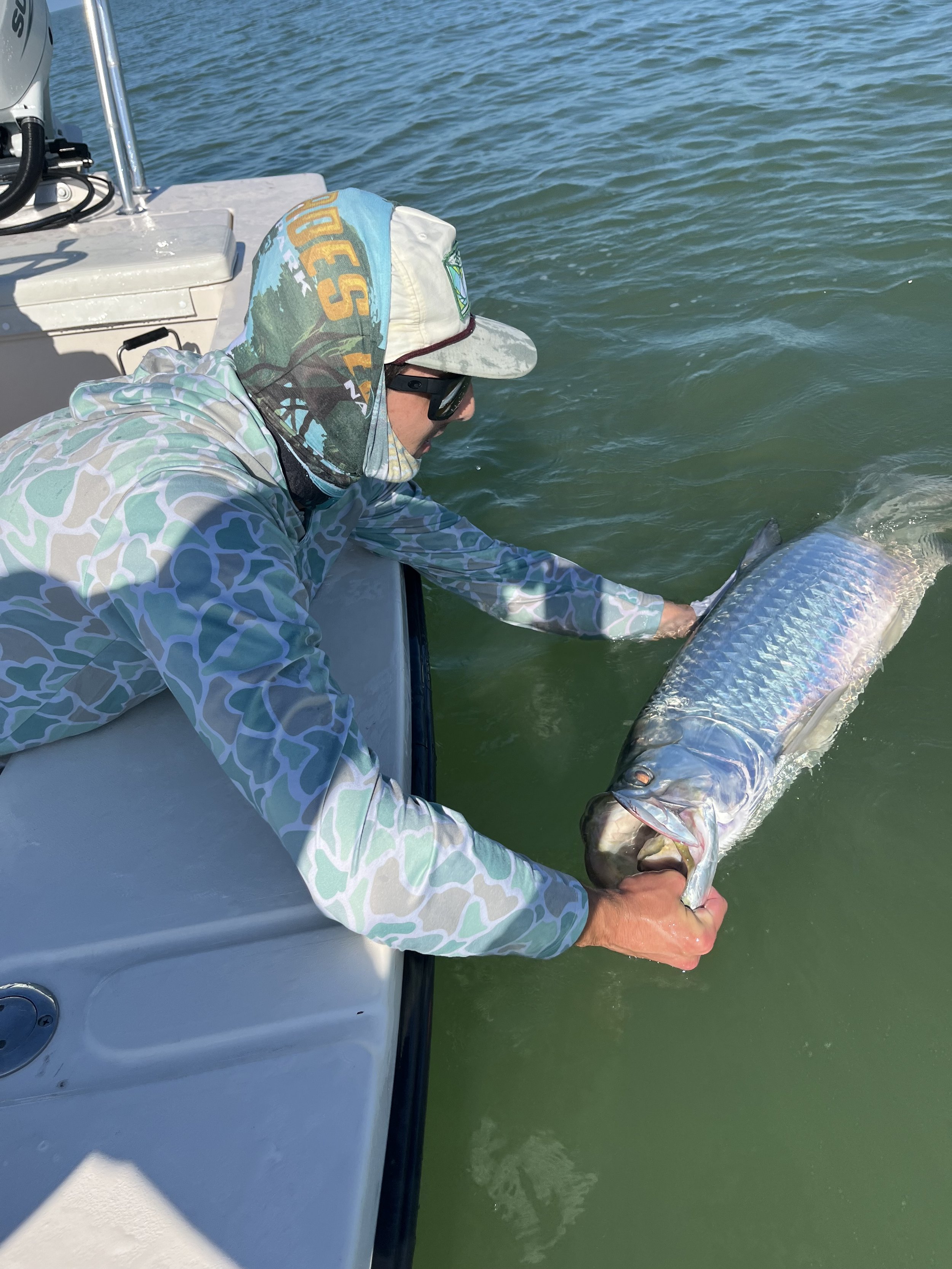 Person in camouflage clothing and sunglasses holding a large fish with shiny scales over the side of a boat in green water.