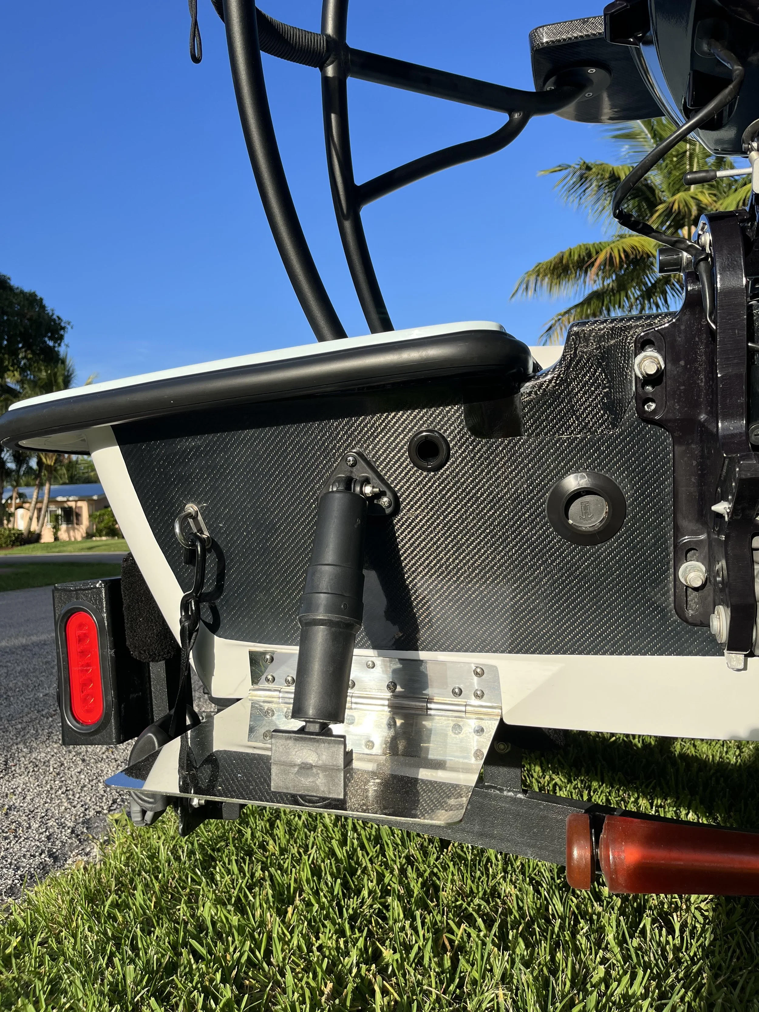 Close-up of a section of a boat with a black carbon fiber panel, hardware, and a handle, under a clear blue sky with palm trees in the background.