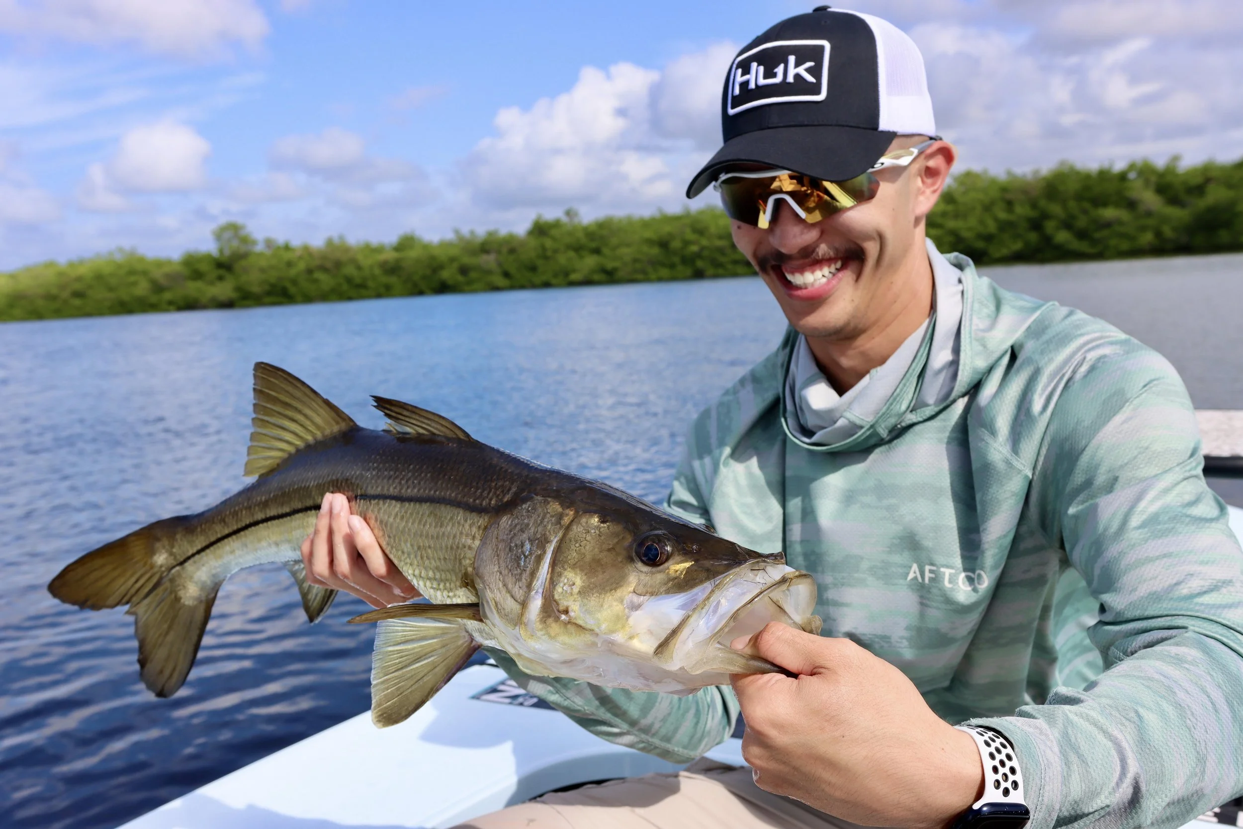A man in sunglasses and a baseball cap holding a fish on a boat by a lake.