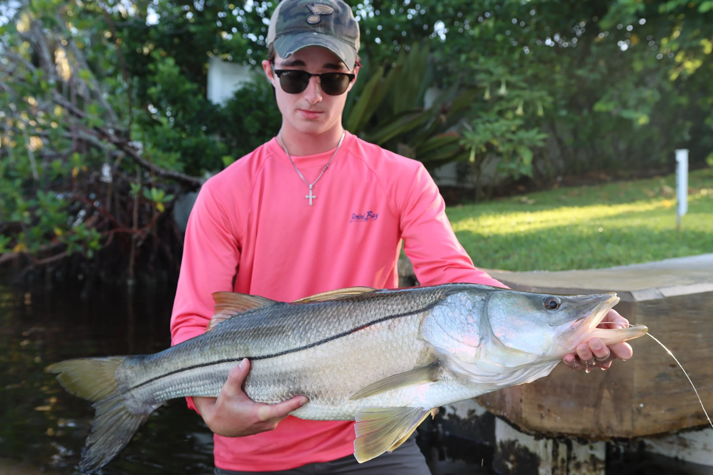 Young man in pink long sleeve shirt, black sunglasses, and a camo hat holding a large fish outdoors near a water body, with green trees and grass in the background.