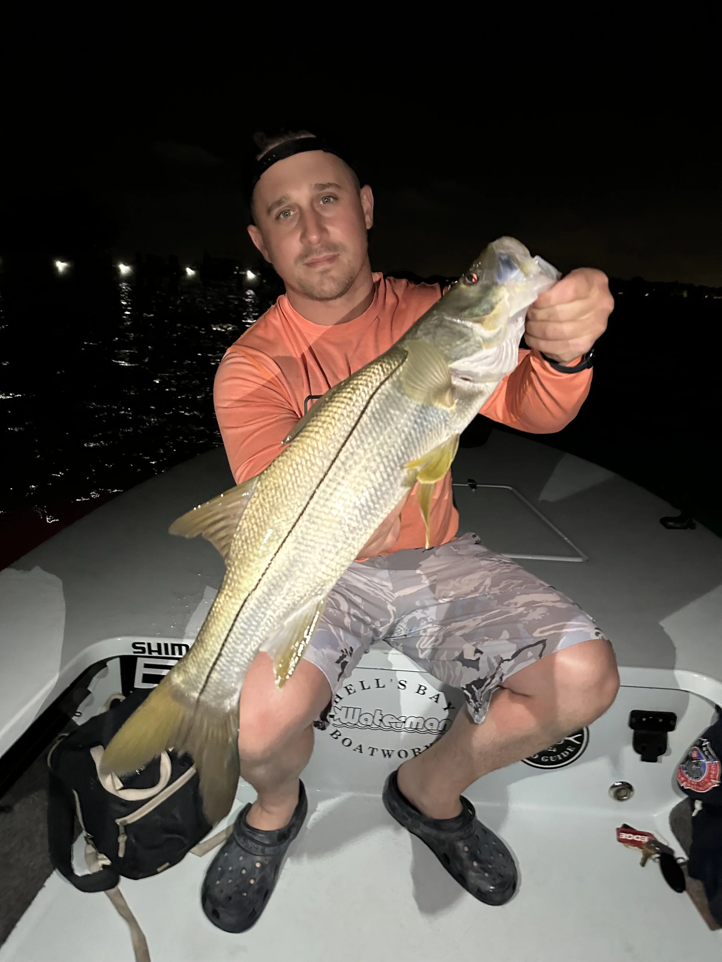 Man in orange shirt and camouflage shorts holding a large fish on a boat at night.