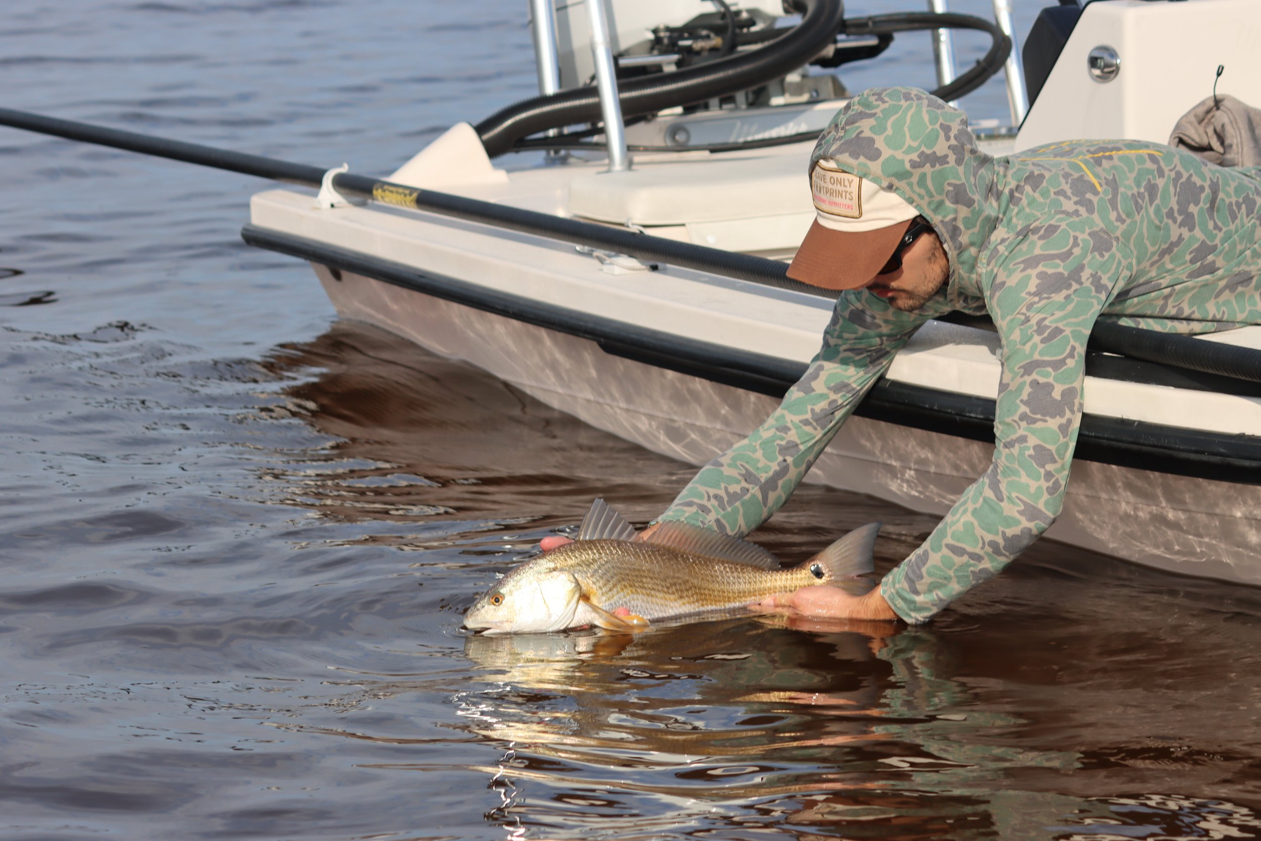 Person in camouflage hoodie and cap releasing a fish back into the water near a boat.