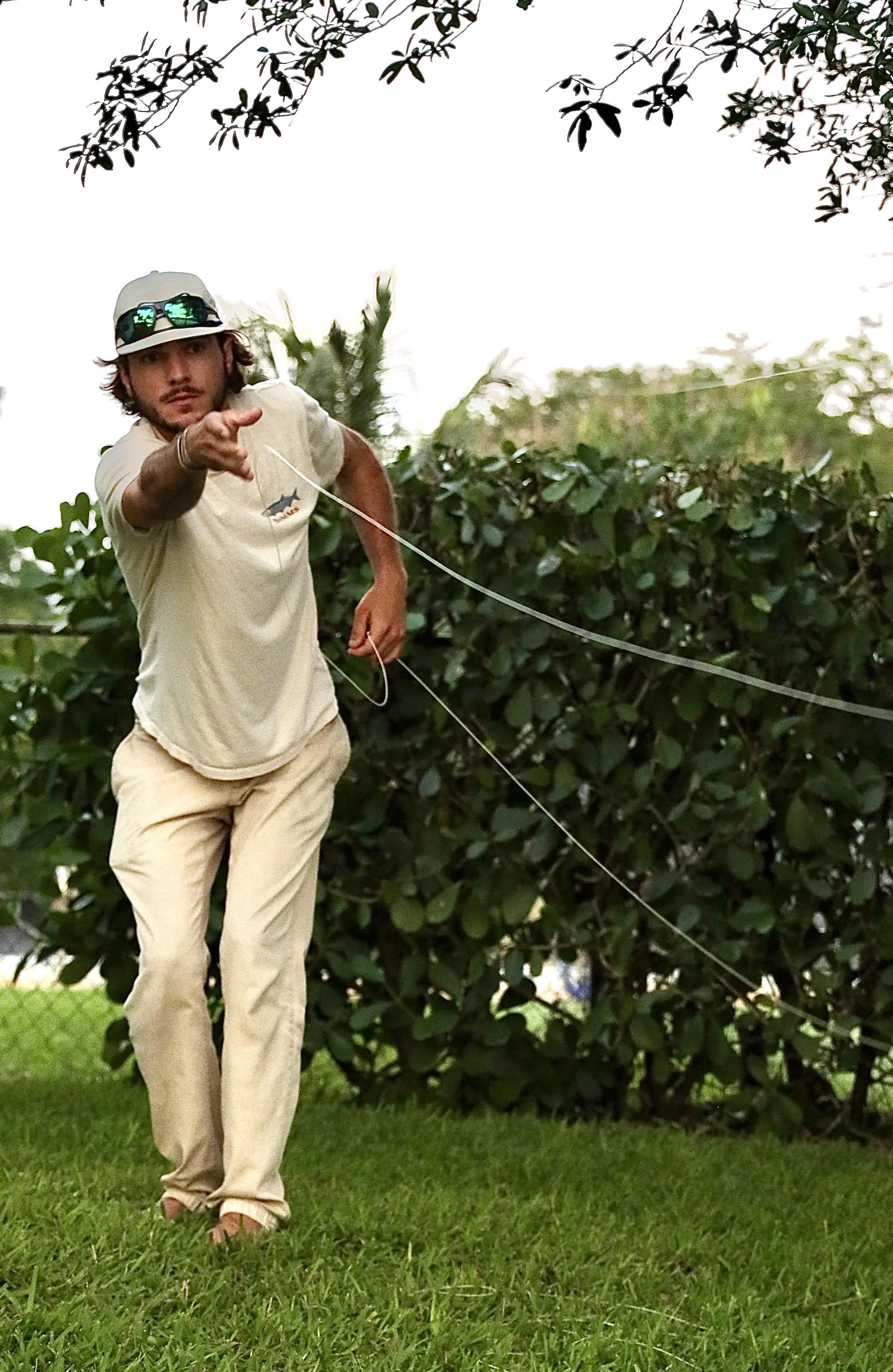 A young man wearing sunglasses, a baseball cap, beige clothing, and flip-flops is pointing at the camera while standing on grass with bushes behind him.