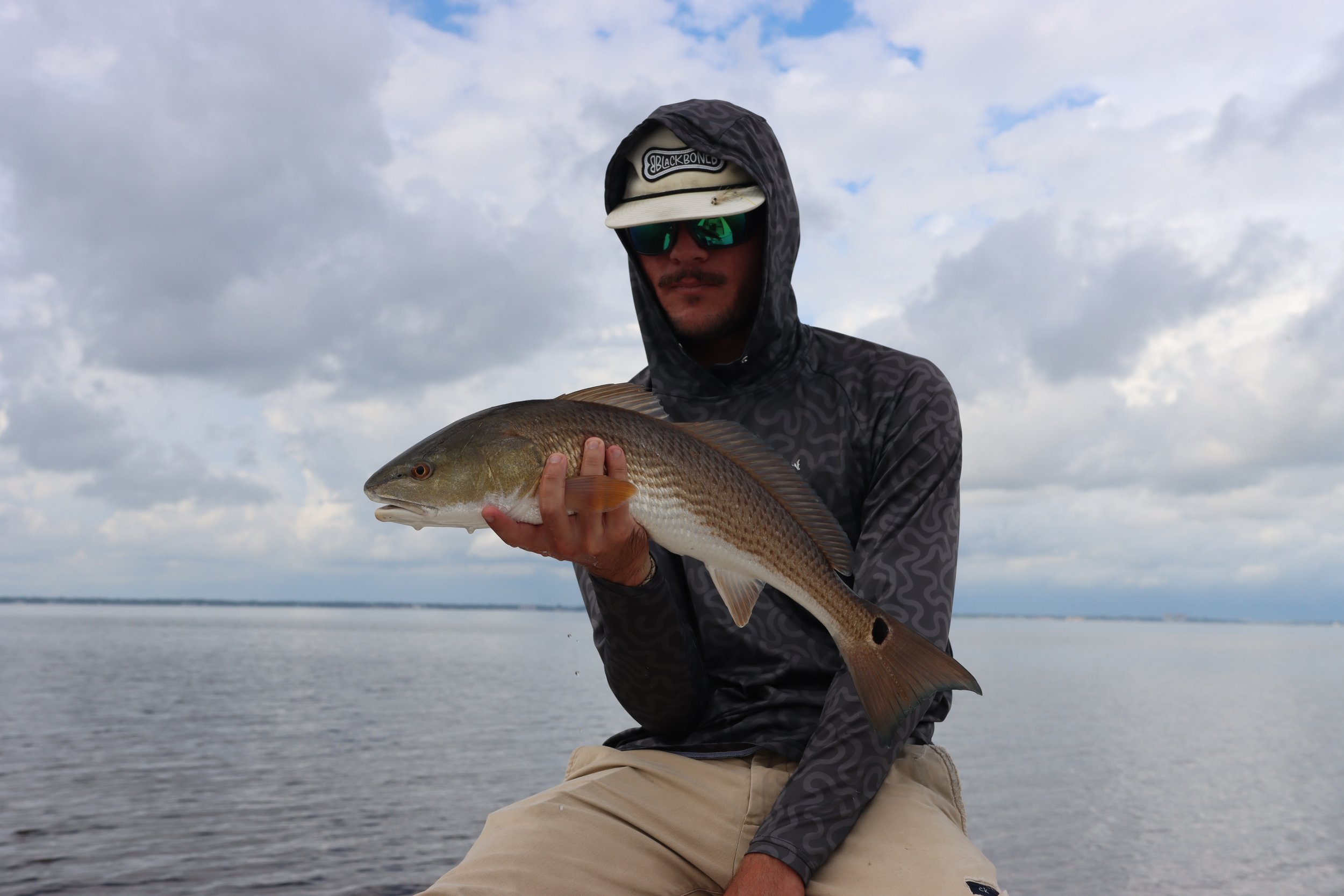 Man in a dark hooded jacket, beige pants, sunglasses, and a hat holding a large fish near a body of water under cloudy sky.