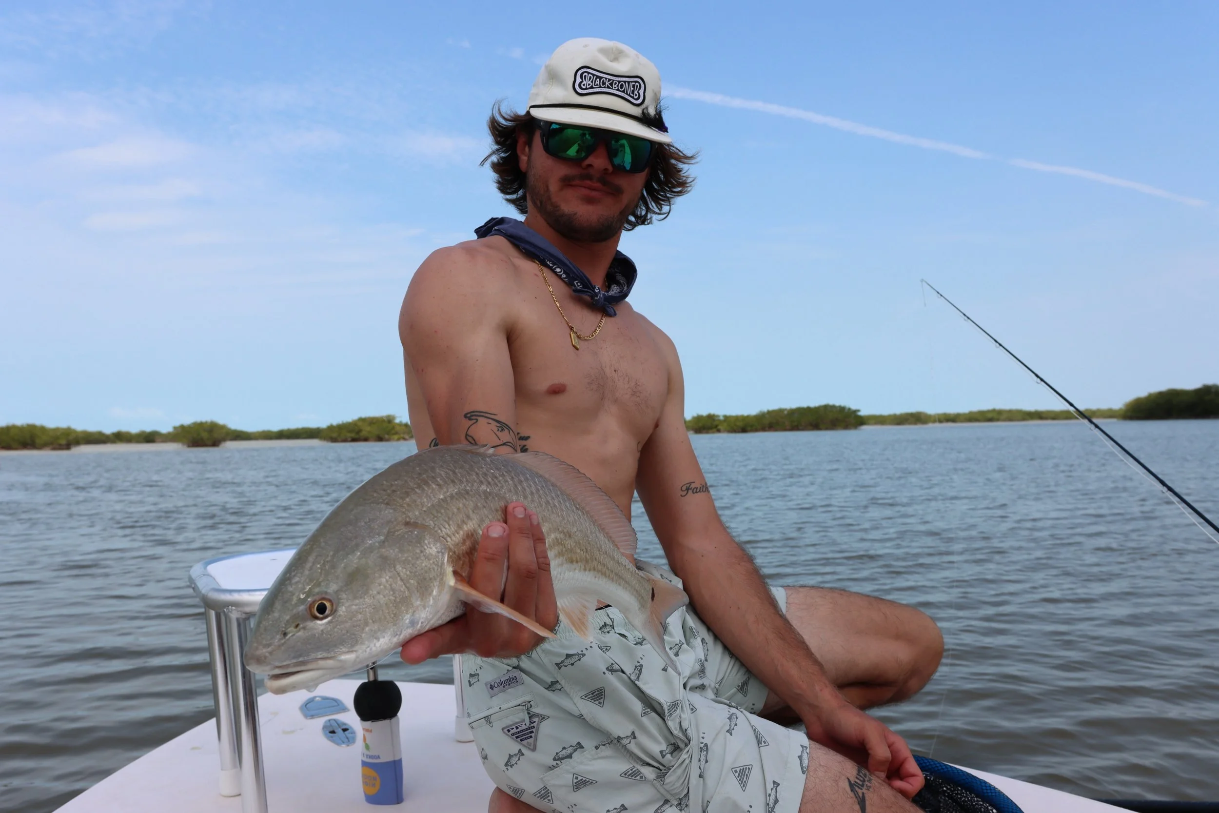 A shirtless man with tattoos, wearing sunglasses, a cap, and patterned shorts, sitting on a boat holding a fish.