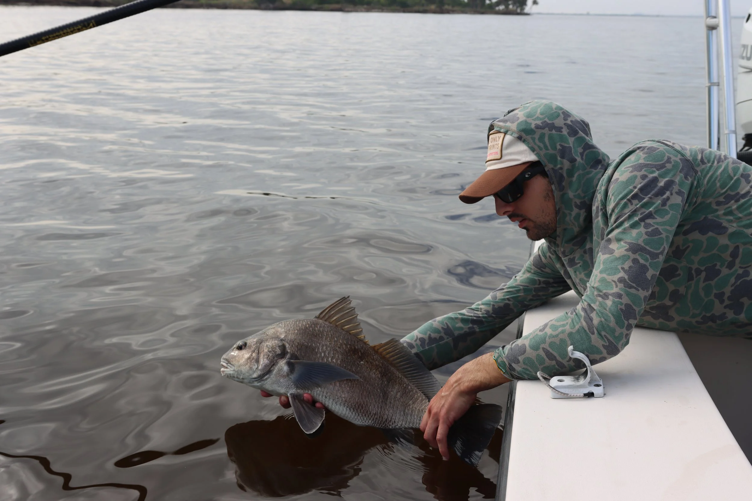 A man in camouflage clothing, sunglasses, and a hat is leaning over the side of a boat, holding a large fish in the water.