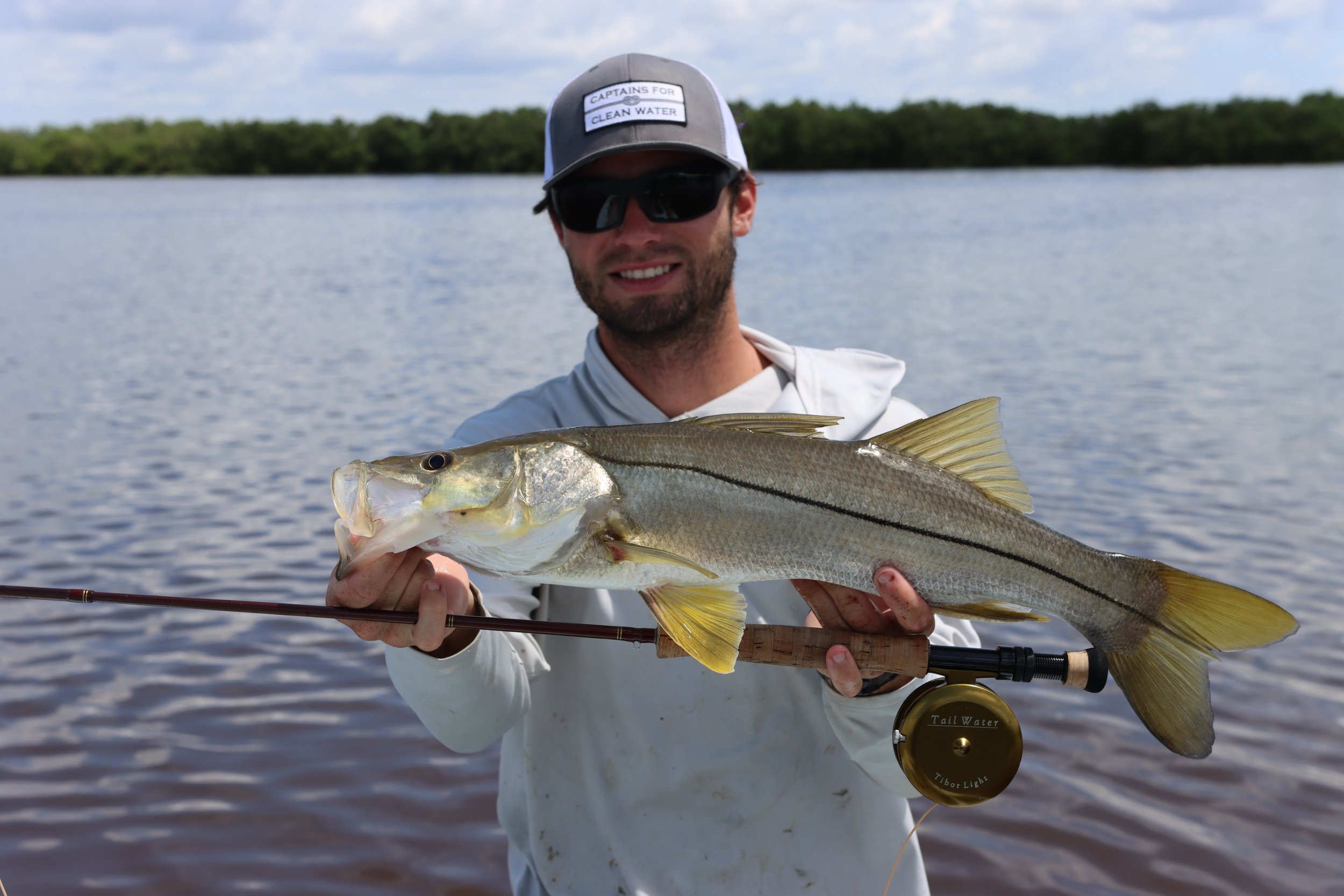 Man holding a large fish over a body of water, wearing sunglasses, a cap, and a white jacket.
