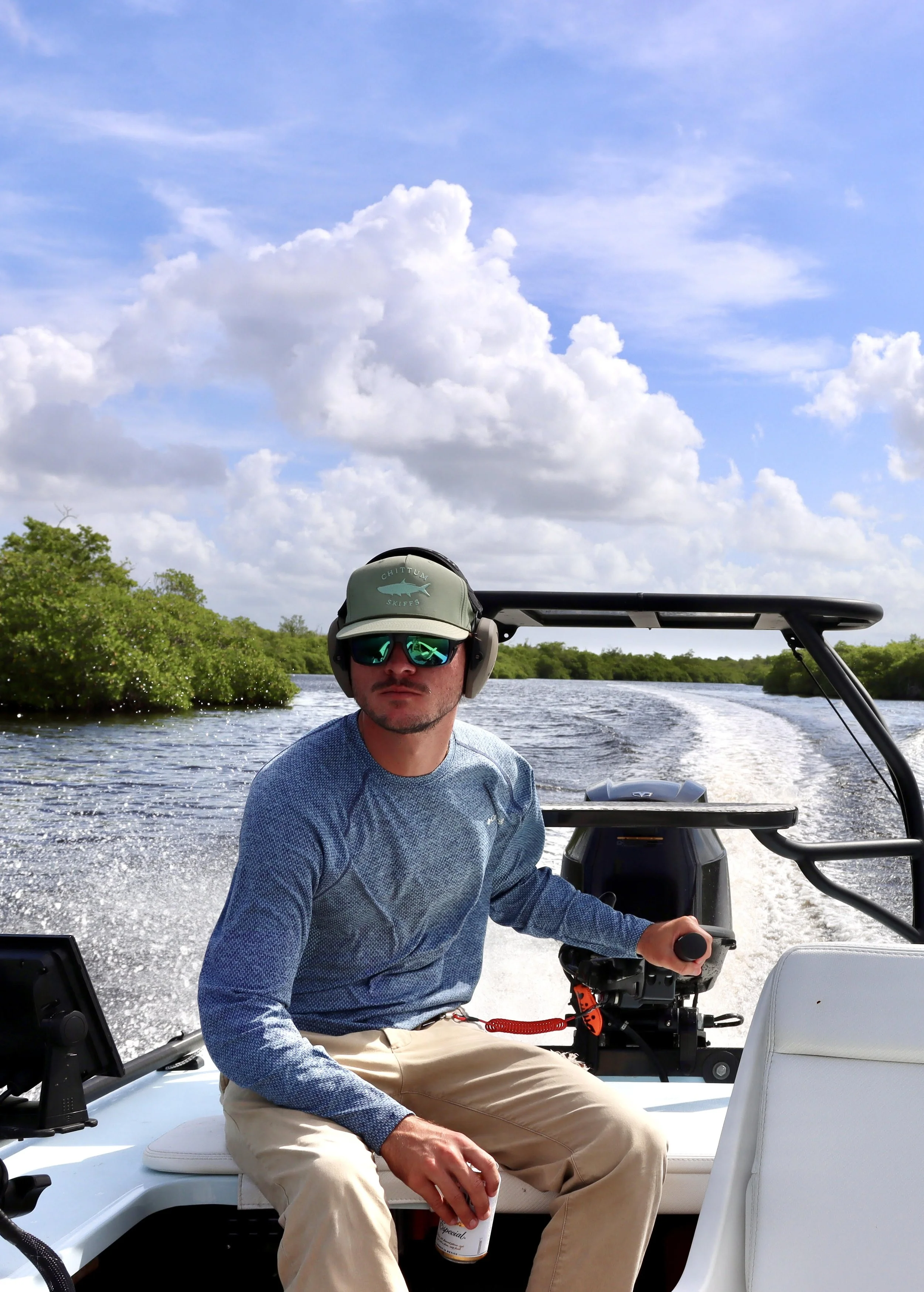 Man in sunglasses and headphones sitting on a boat steering a motorboat on a river with greenery and cloudy sky in the background.
