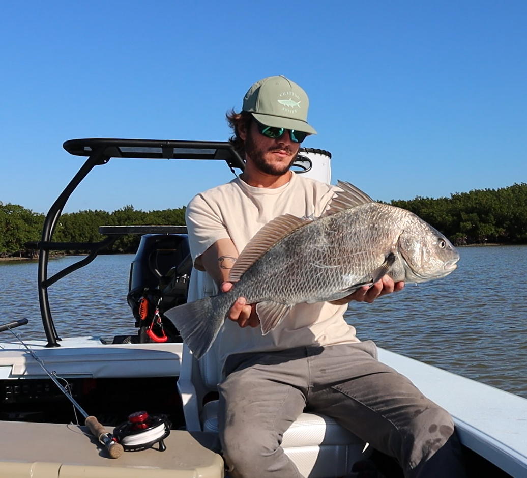 Man sitting on a boat holding a large fish he caught, with water and trees in the background.