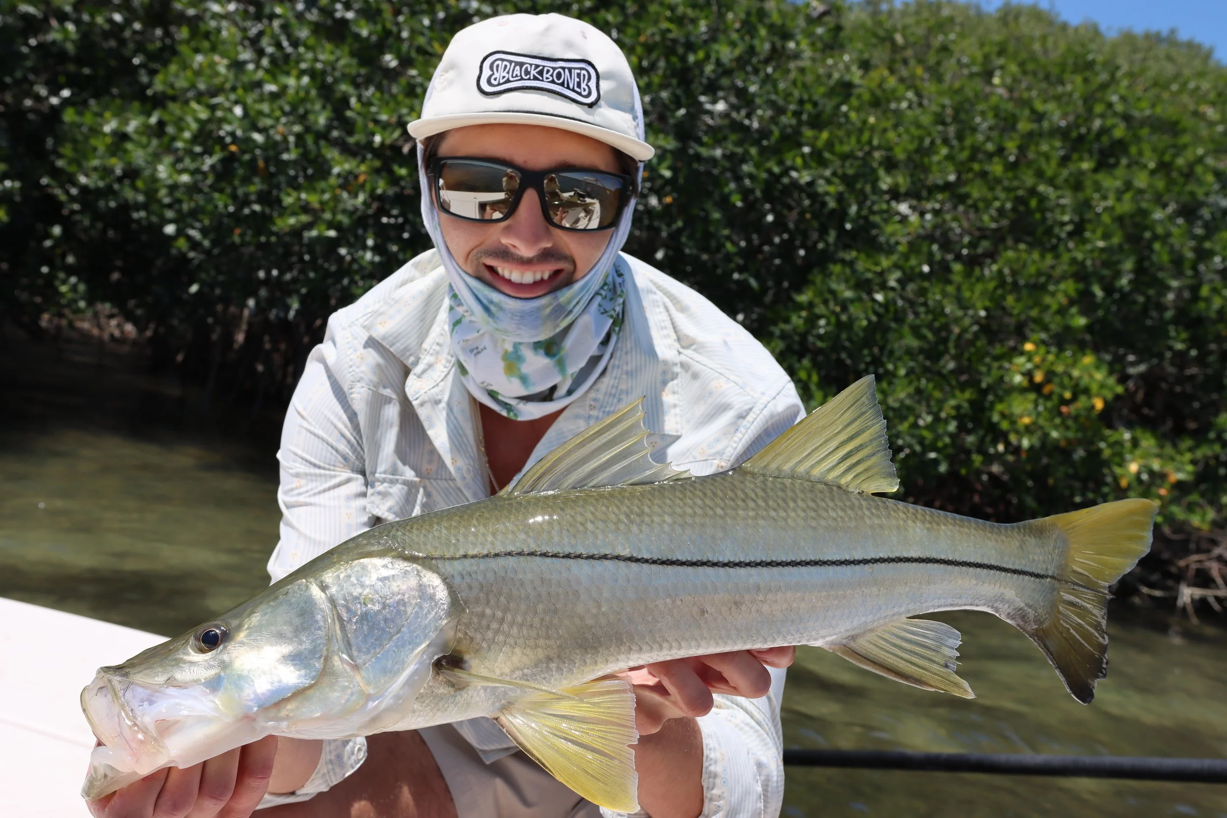 Man wearing sunglasses, a white cap with the word 'Blackbone', and a floral neck gaiter holding a large fish, possibly a snook, near a body of water with greenery in the background.