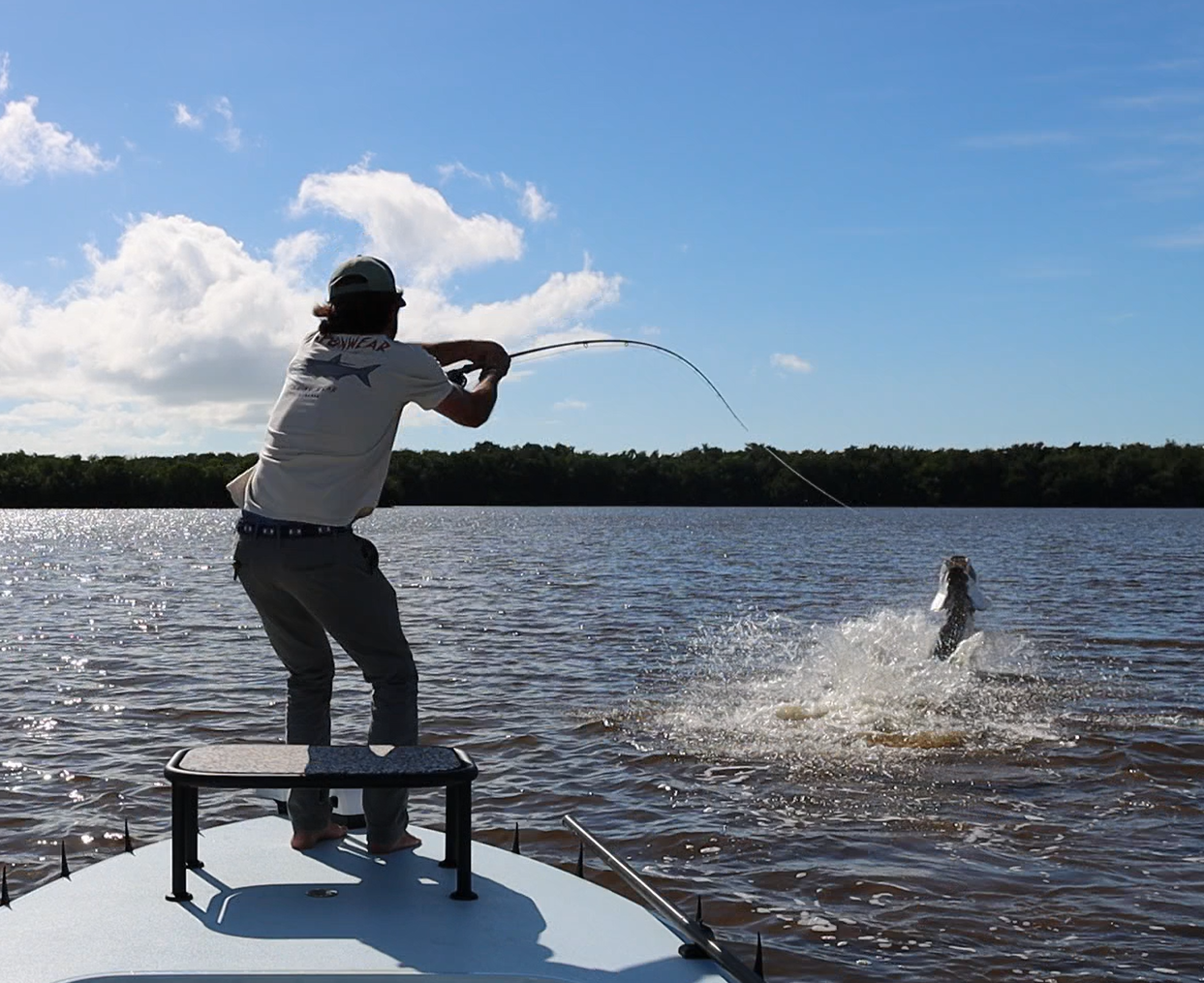 Person fishing from a boat, reeling in a large fish in a lake with a blue sky and some clouds.