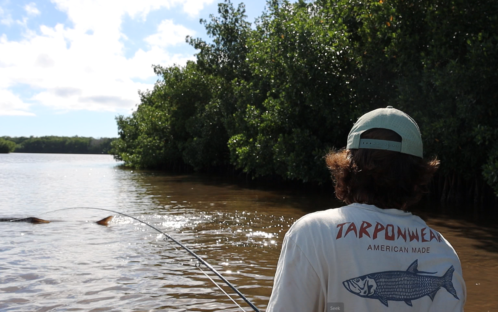 Person with curly hair fishing in a river, wearing a light-colored cap and a T-shirt with a fish illustration and the word "TARPONWEAR" on the back, surrounded by lush green trees along the riverbank under a partly cloudy sky.