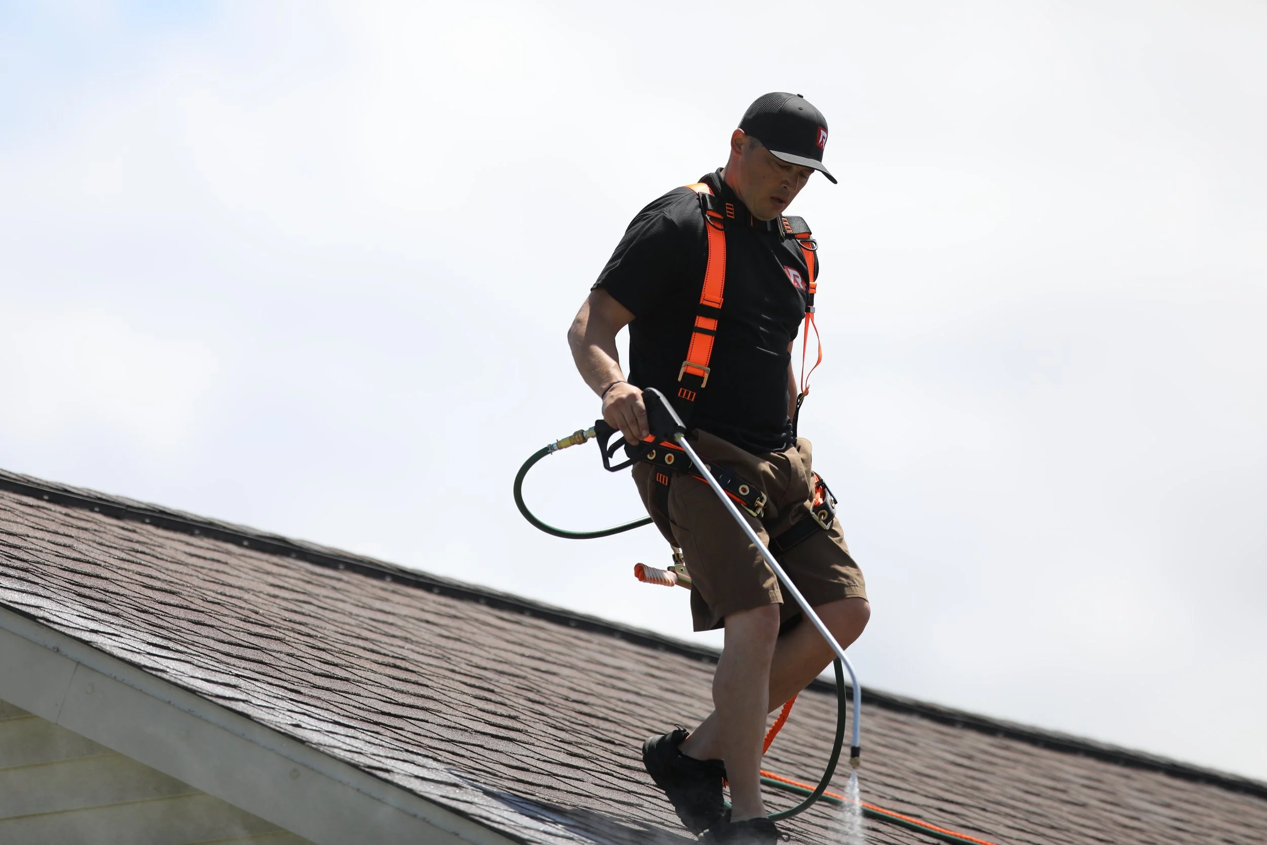 A man working on a roof with a sprayer, wearing safety gear including a harness, cap, and shorts.