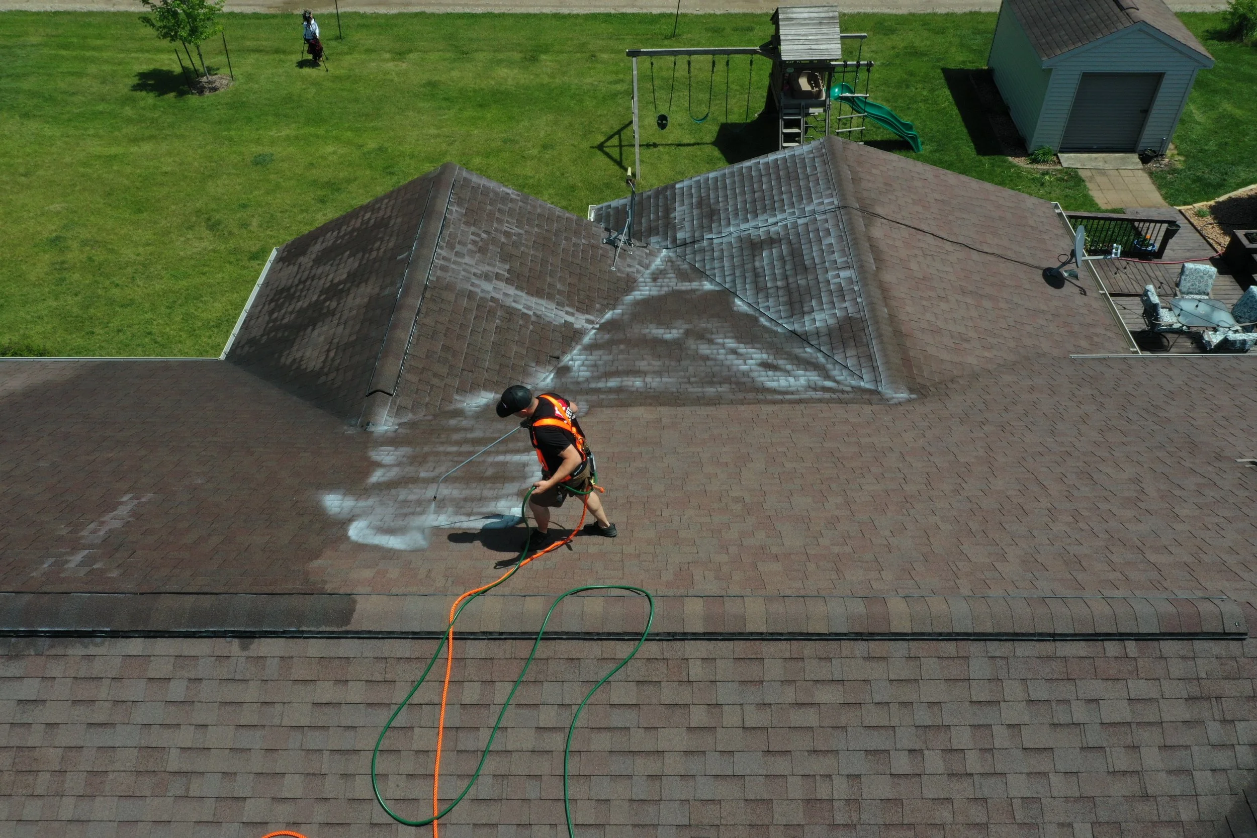 A person wearing a black helmet, safety vest, and harness power washing a residential roof with a spray wand connected to a hose, creating a white streak on the shingles.