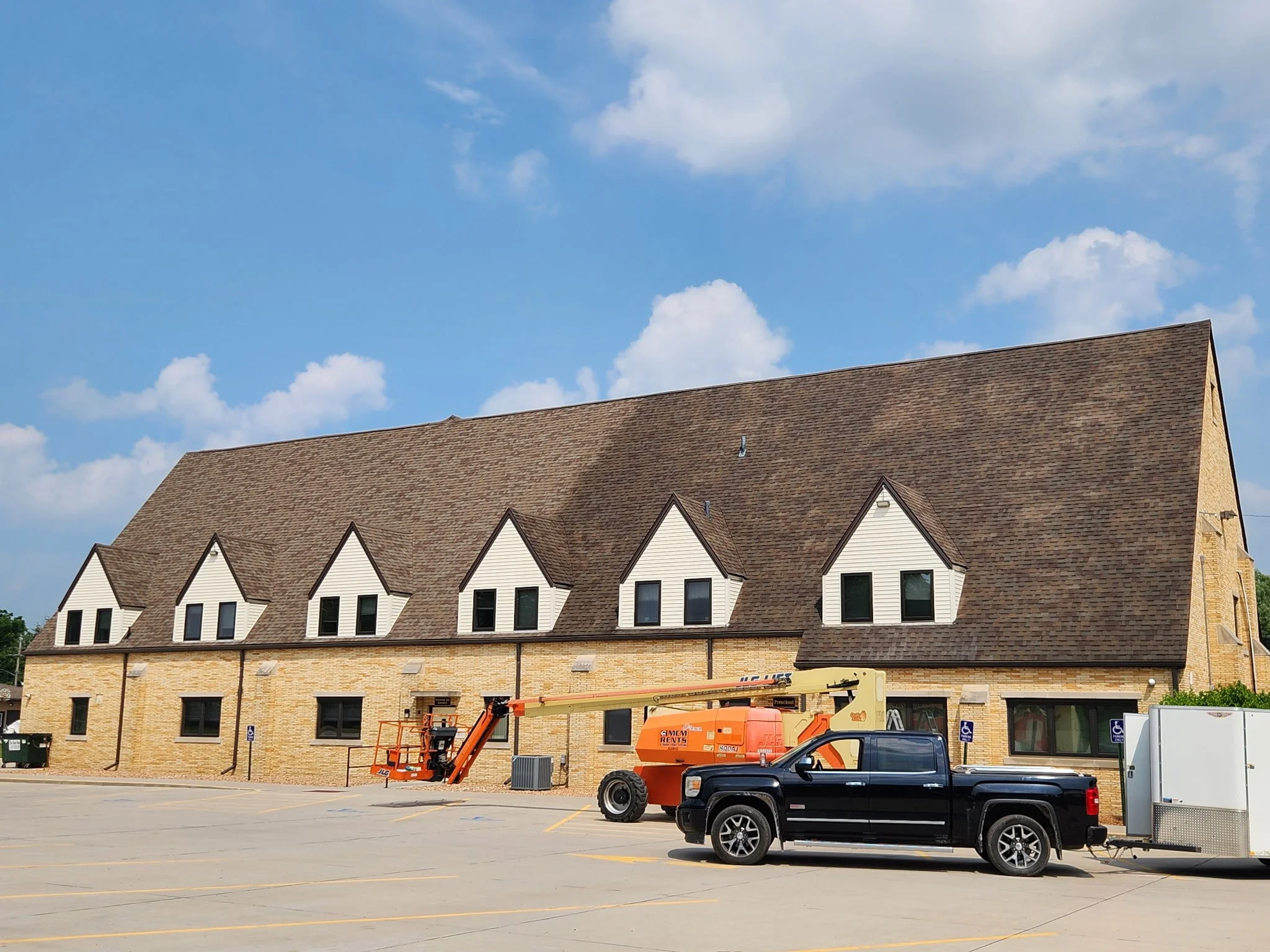 A multi-story building with a sloped roof and dormer windows, construction equipment, and a parked black truck in a nearly empty parking lot under a partly cloudy sky.
