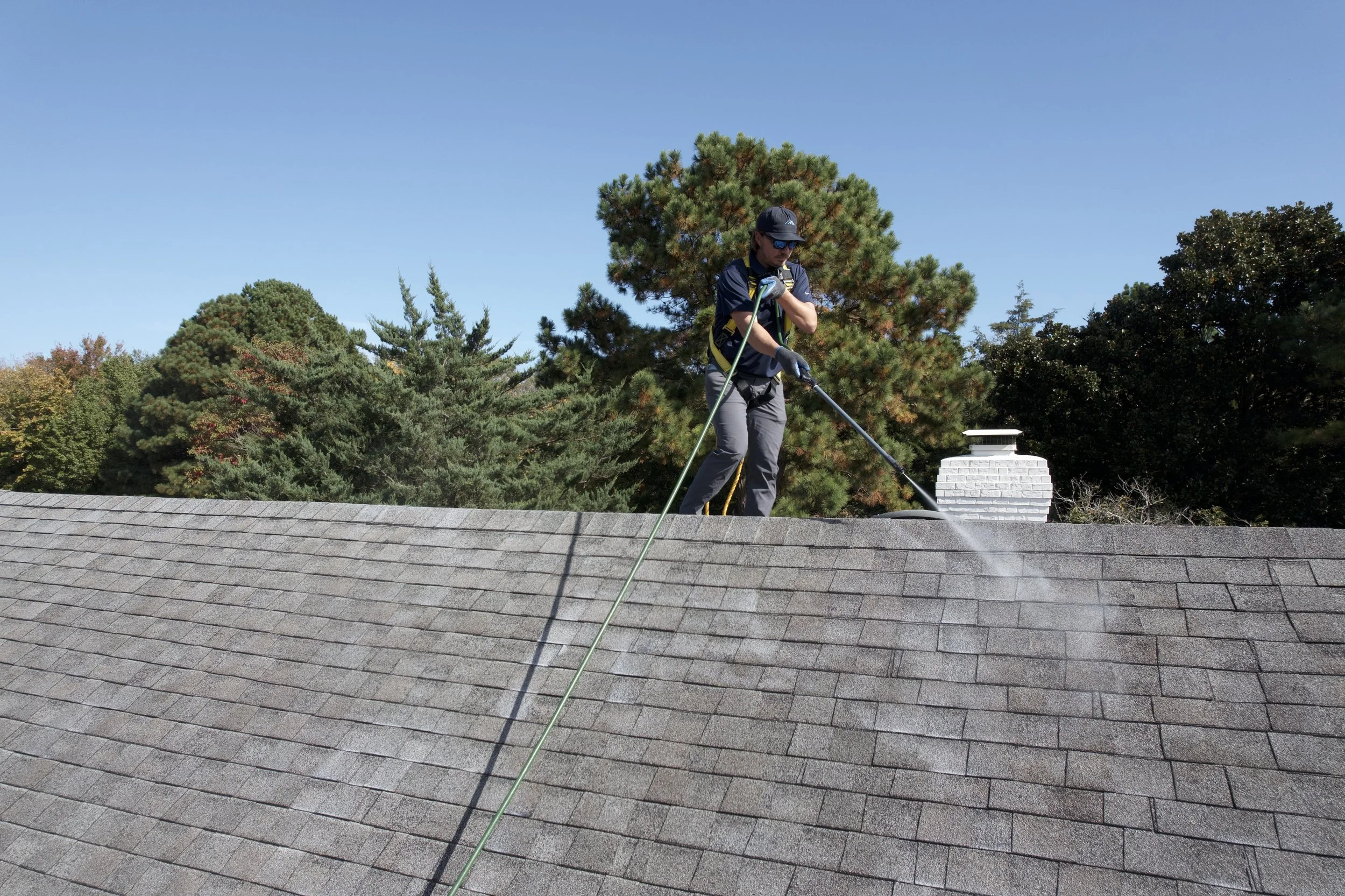 A person wearing sunglasses, a baseball cap, and gloves is pressure washing a roof with a high-pressure spray, standing on the shingles. There are green trees and a blue sky in the background.