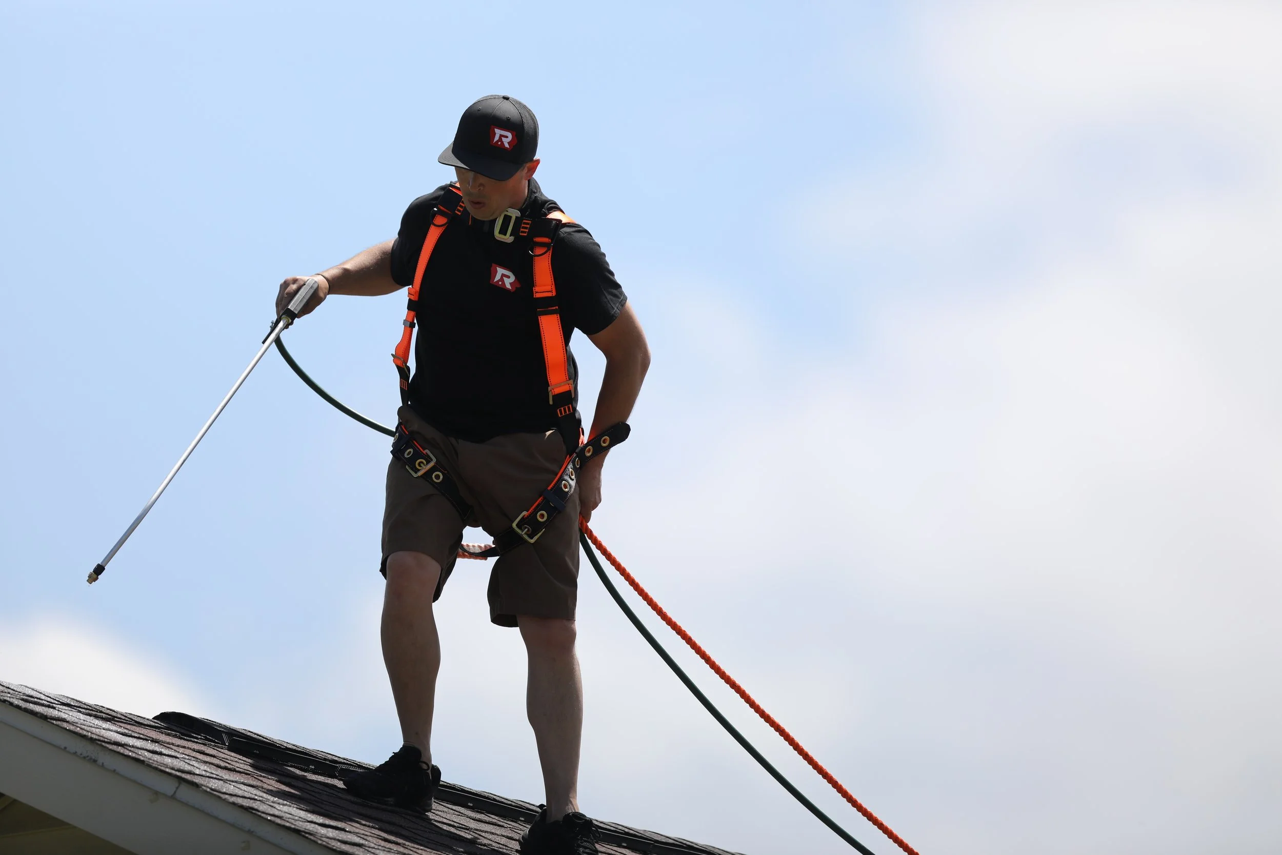 A man standing on a roof, holding a long metal tool, wearing safety gear including a harness, helmet, and a backpack, against a partly cloudy sky.