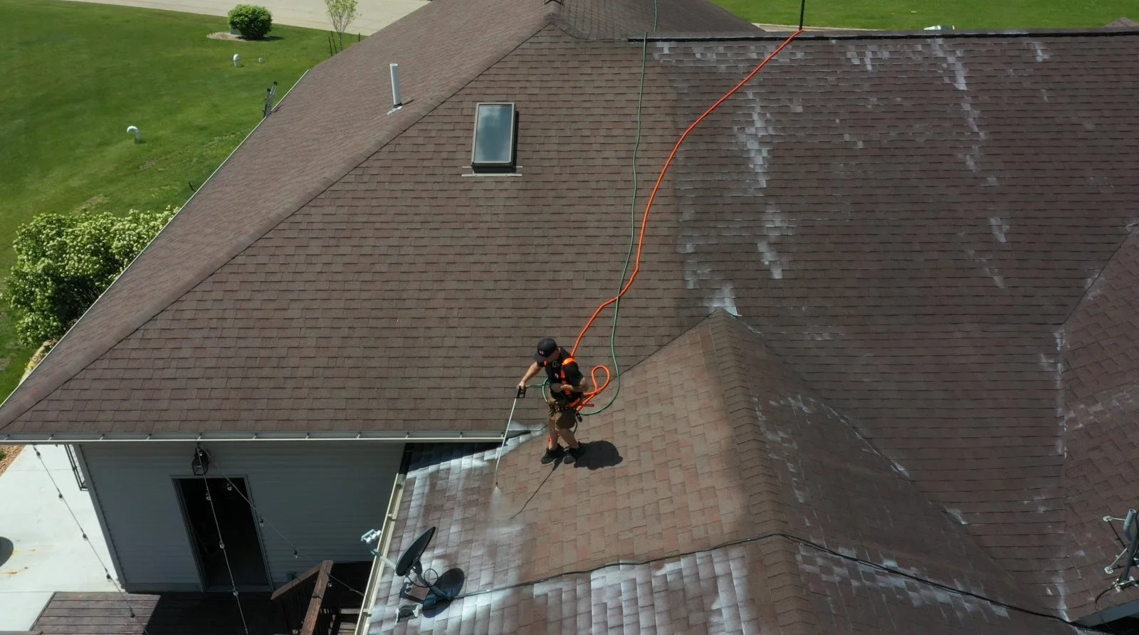 A person cleaning dirt and moss off a brown shingled roof with a pressure washer, with green grass and trees in the background.