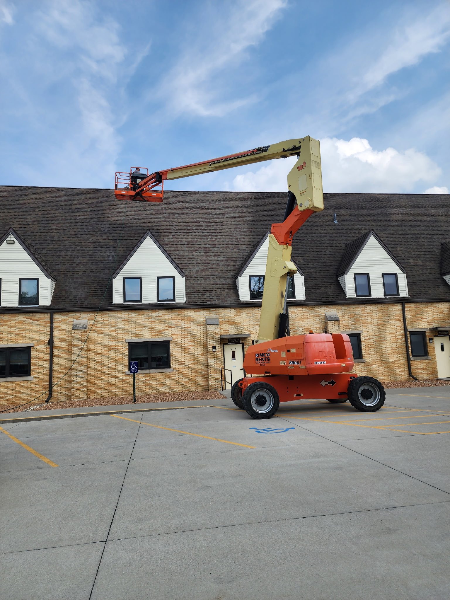A cherry picker with a worker on the lift is in a parking lot next to a brick building with white dormer windows and a steep roof. The parking lot has yellow lines and a handicapped parking space.