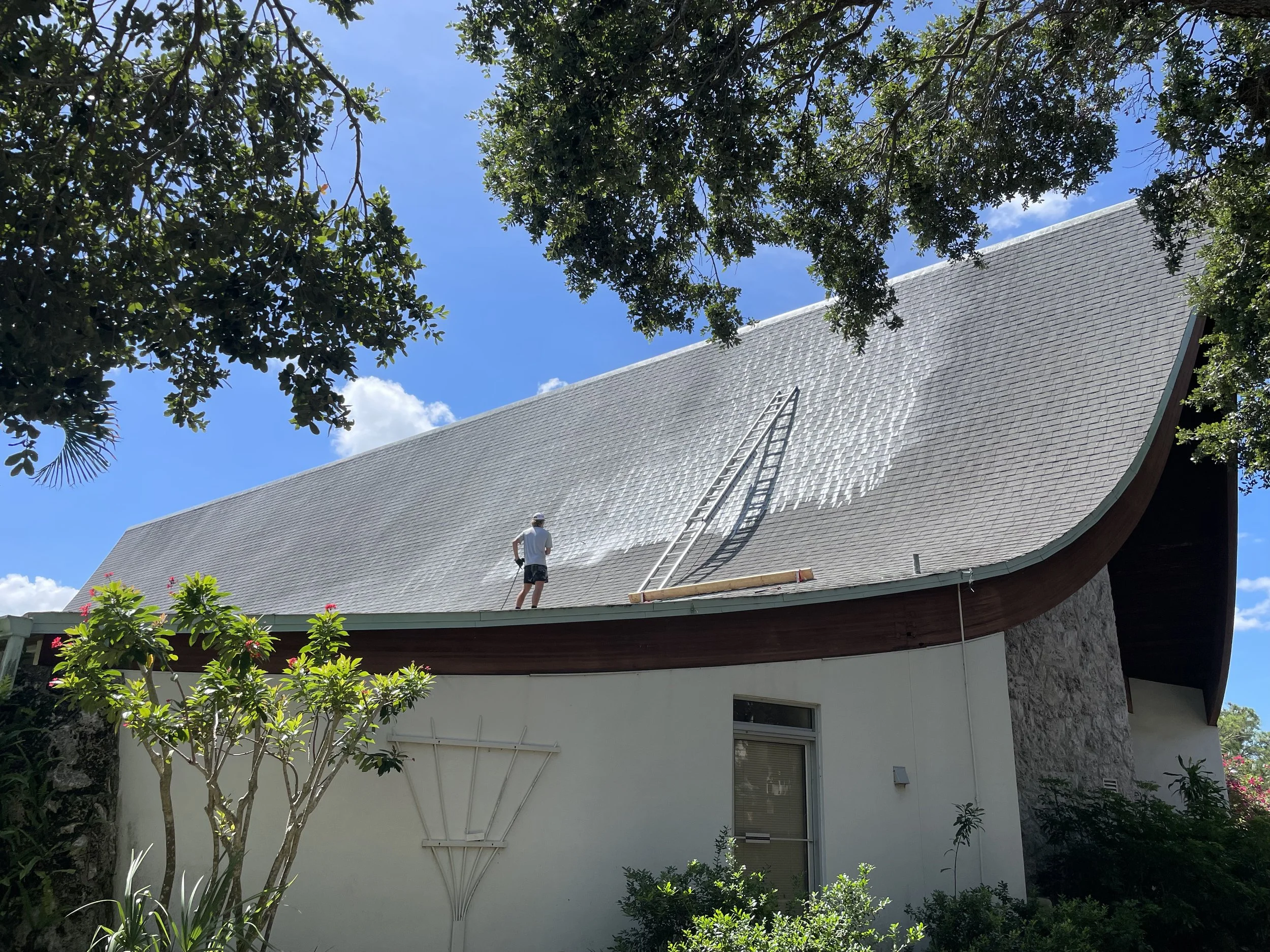 A man standing on a roof, painting or cleaning the large, curved, shingle roof of a modern house with a ladder leaning against it, surrounded by lush green trees and a partly cloudy blue sky.