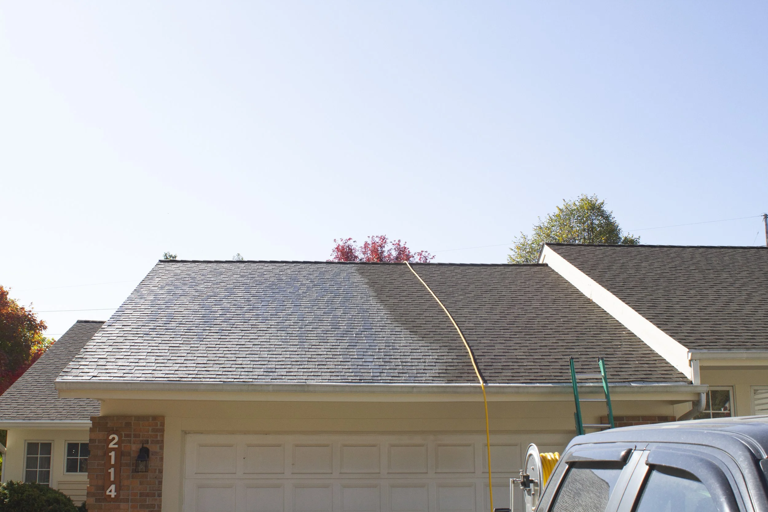 Cleaning or pressure washing a house roof with a yellow hose, vehicle and ladder nearby, number 2114 on the house