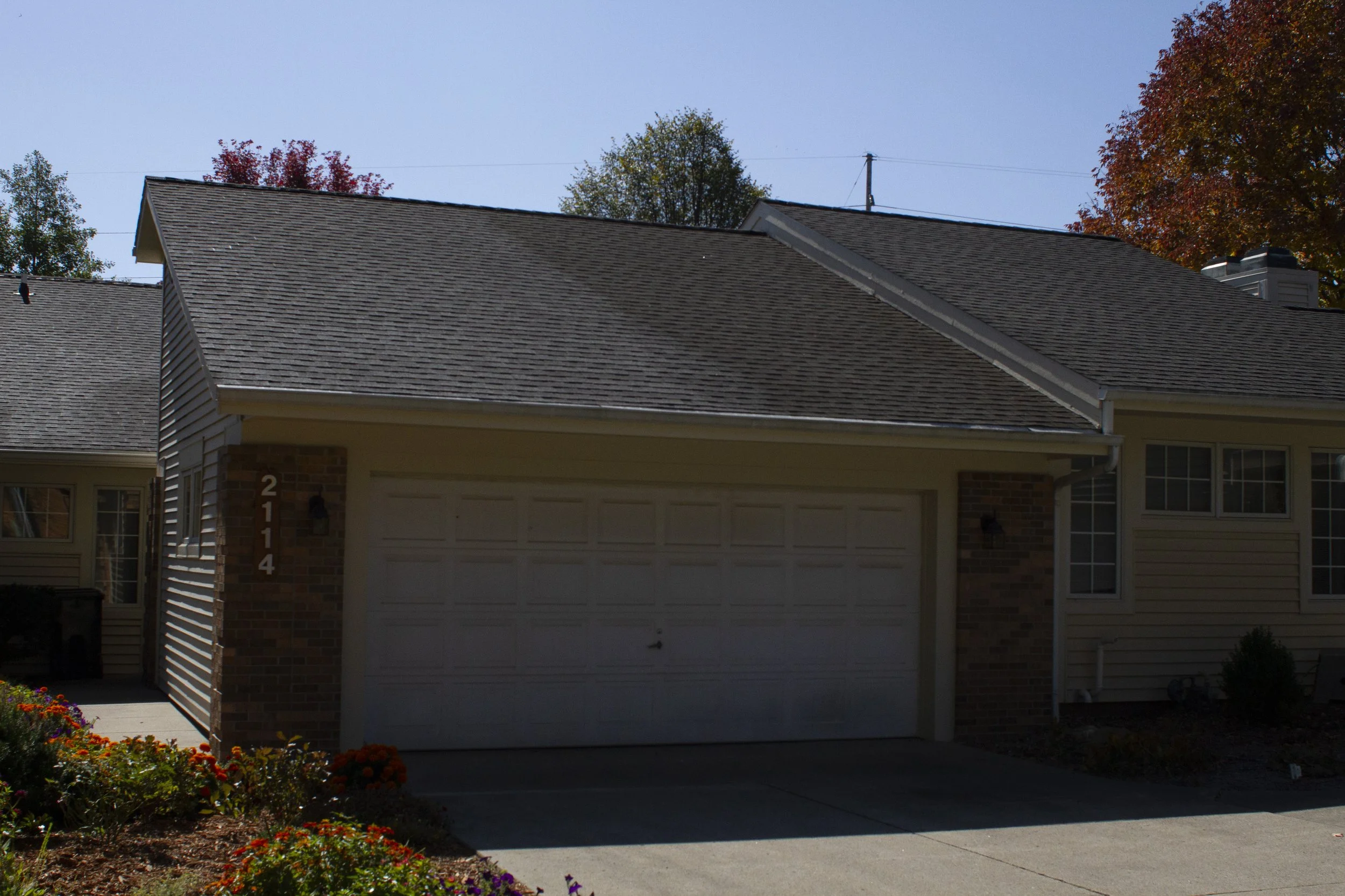 Front view of a house with a closed garage door, brick and siding exterior, house number 2114 on the brick wall, and some colorful flowers in the garden in front.