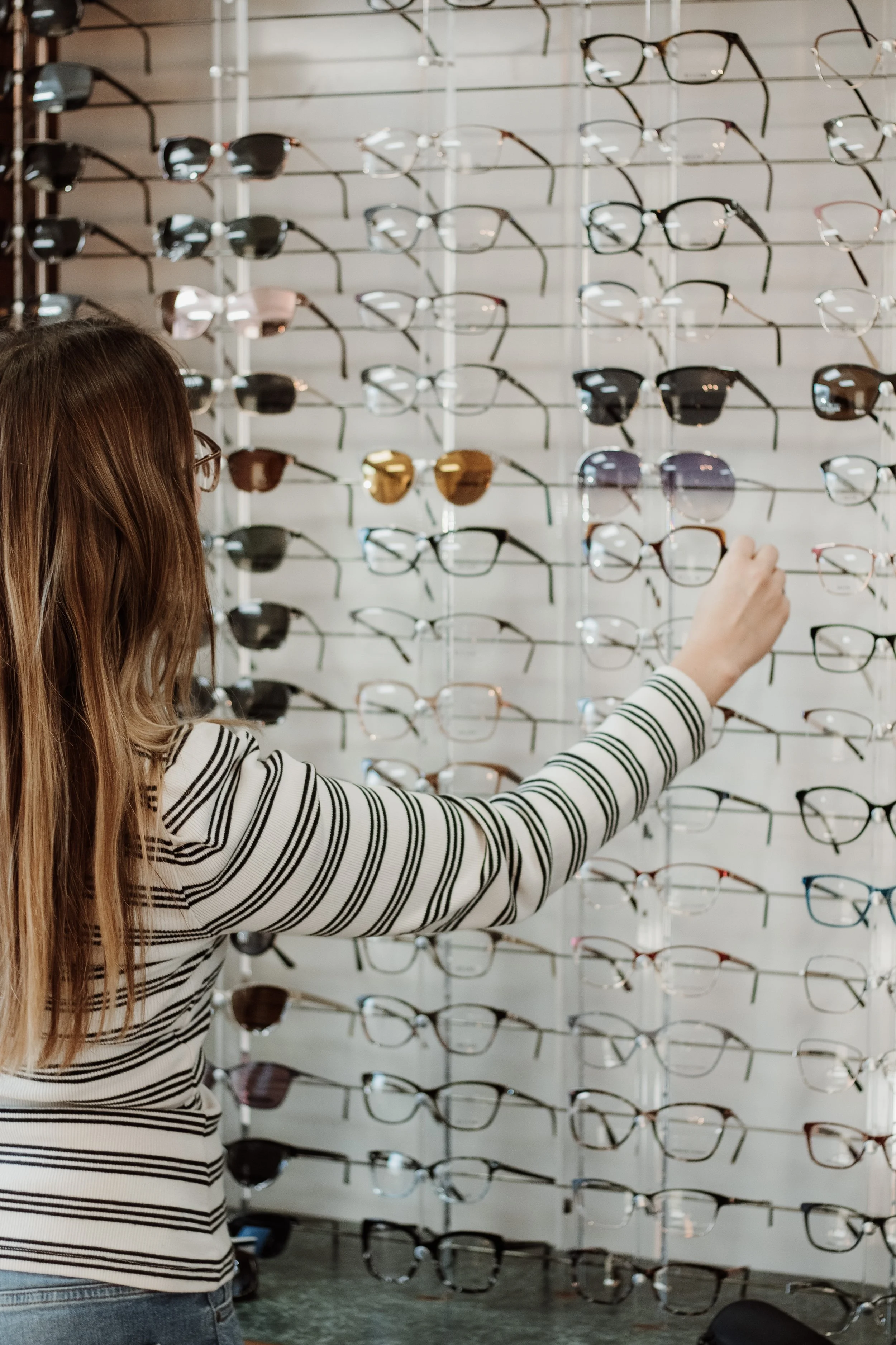 A woman with long hair and glasses shopping for glasses at an eyewear store, selecting a frame from a wall of various eyeglasses.