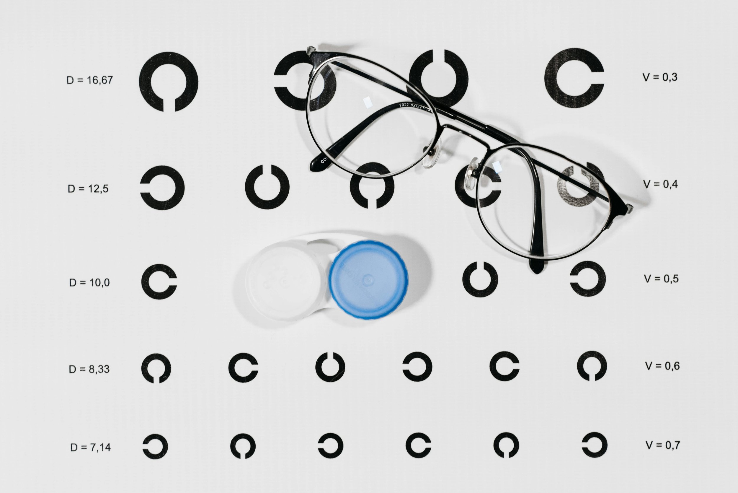 A visual acuity test chart with rings of different sizes and glasses placed on top. There are two contact lens cases with blue and white lids in the center.