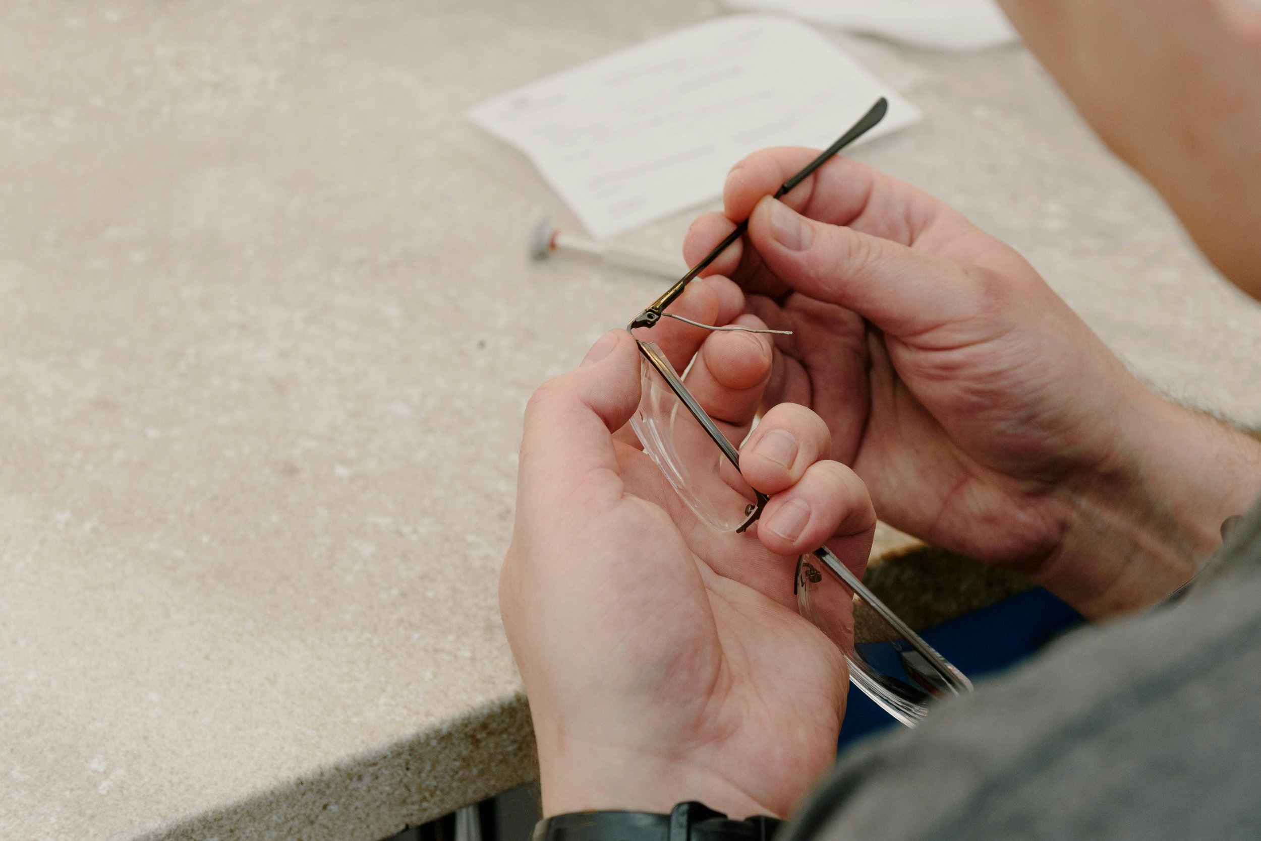 Person repairing eyeglasses with small screwdriver on light-colored countertop, with a blurred paper or instruction sheet in the background.