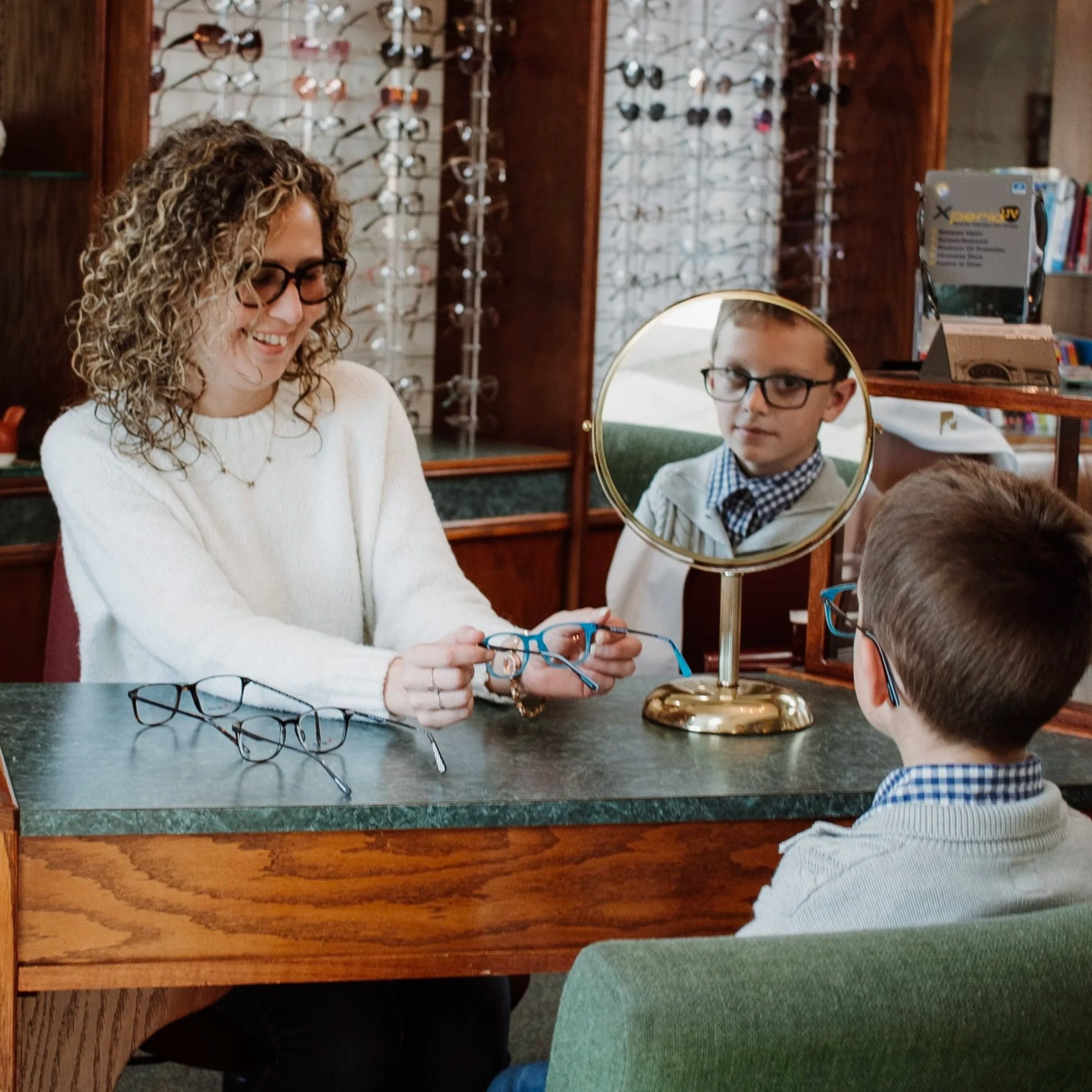 A woman is assisting a young boy at an optometry shop, showing him a pair of glasses. A boy with glasses is looking into a mirror on the counter, while another boy with glasses is sitting across from them. Several pairs of glasses are on the counter.