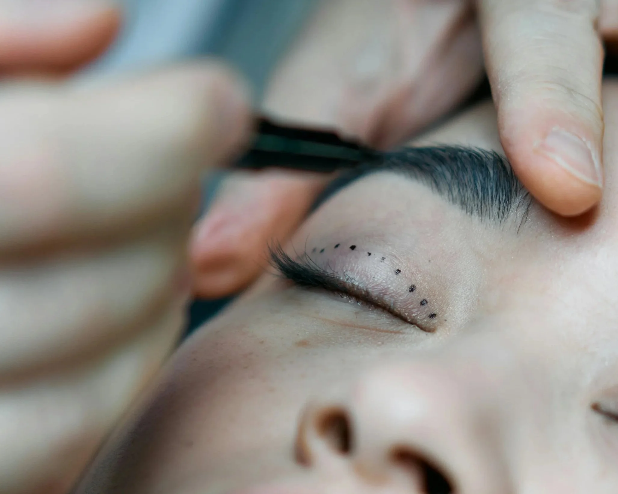 A person receiving makeup or eyelash extension application, with a close-up of the eye and technician's hand holding tools.
