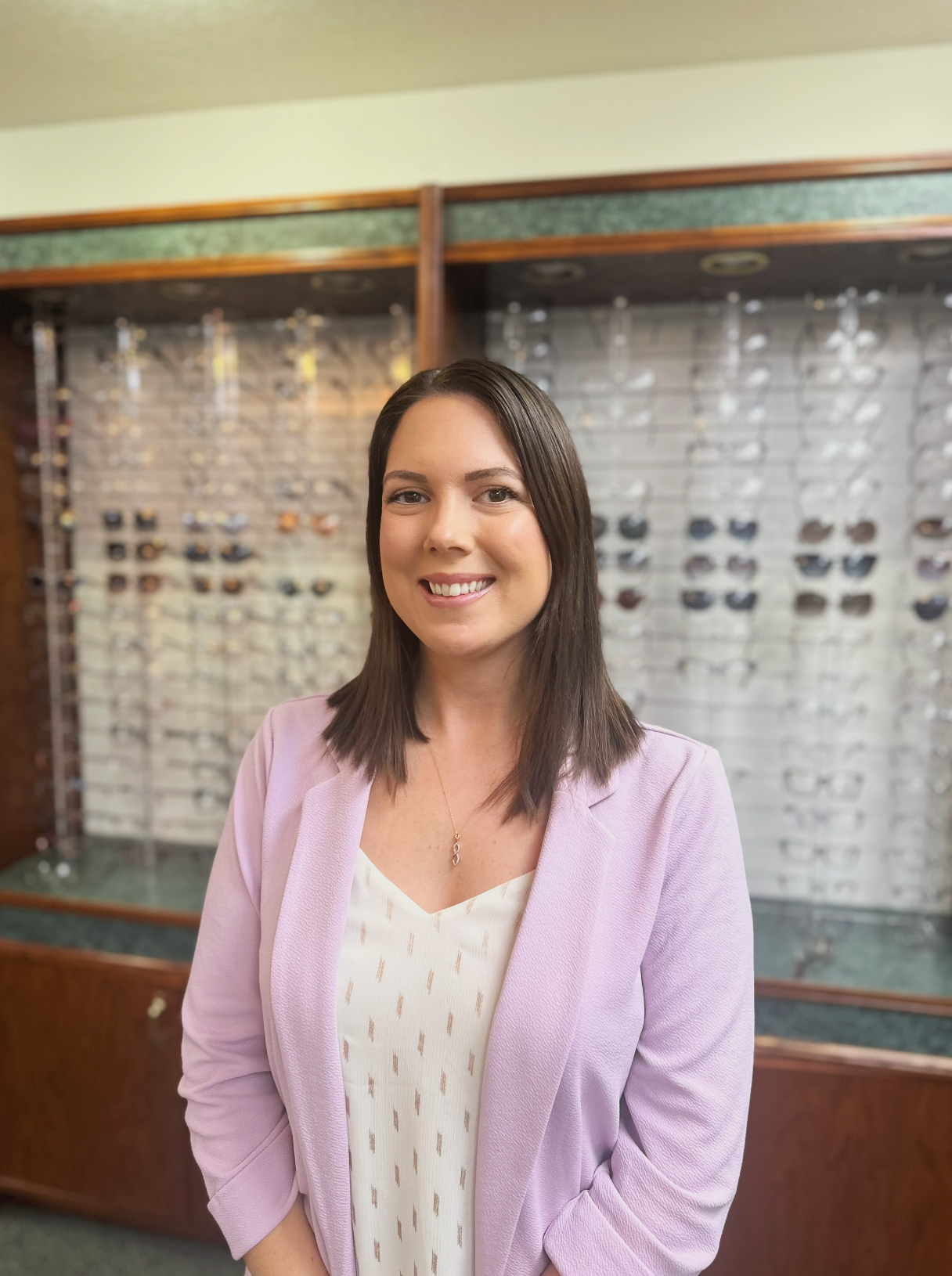 Smiling woman with shoulder-length brown hair wearing a light pink blazer and white blouse, standing in front of an eye glasses display in an eyewear store.