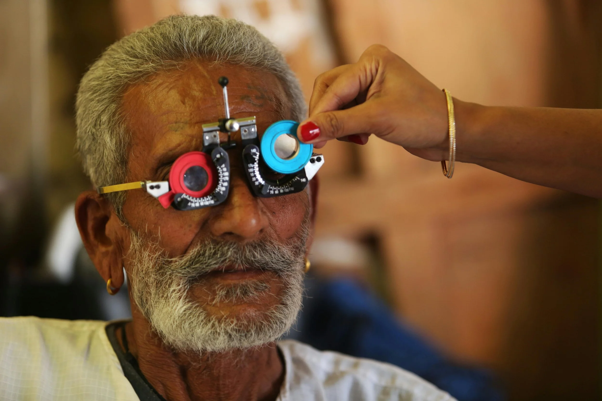 An elderly man with gray hair and a beard getting an eye exam with a phoropter, while a woman adjusts the instrument for his eye measurement.